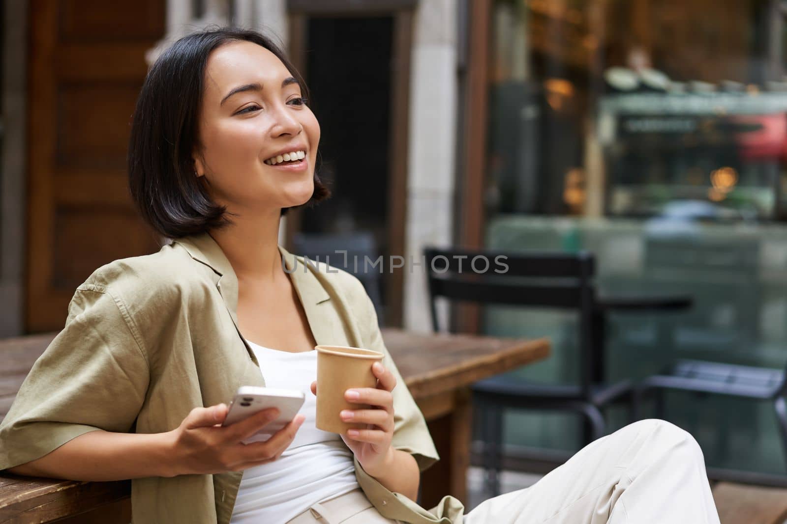 Stylish urban girl sits with her phone in cafe, drinks coffee and chatting, browsing social media on smartphone.