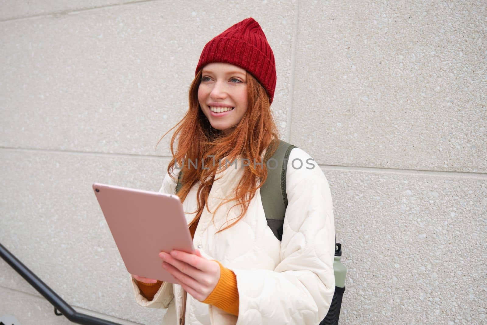 Young redhead girl, walks with digital tablet around city, stands on street with backpack and gadget, looks at online map, searches for a place in internet.