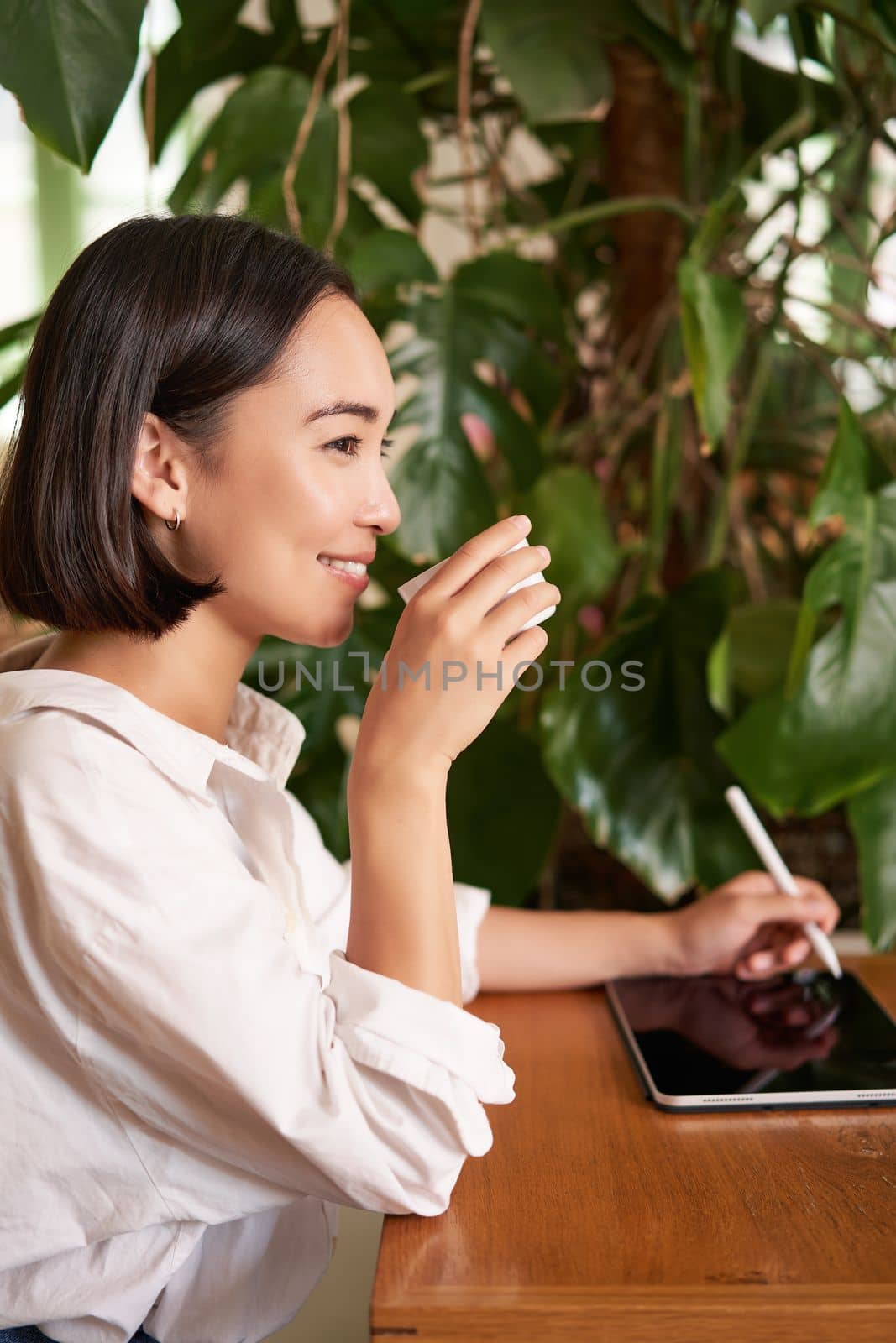 Beautiful asian girl sitting in cafe with cup of coffee, drawing on digital tablet with graphic pen, doddling and making scatches.