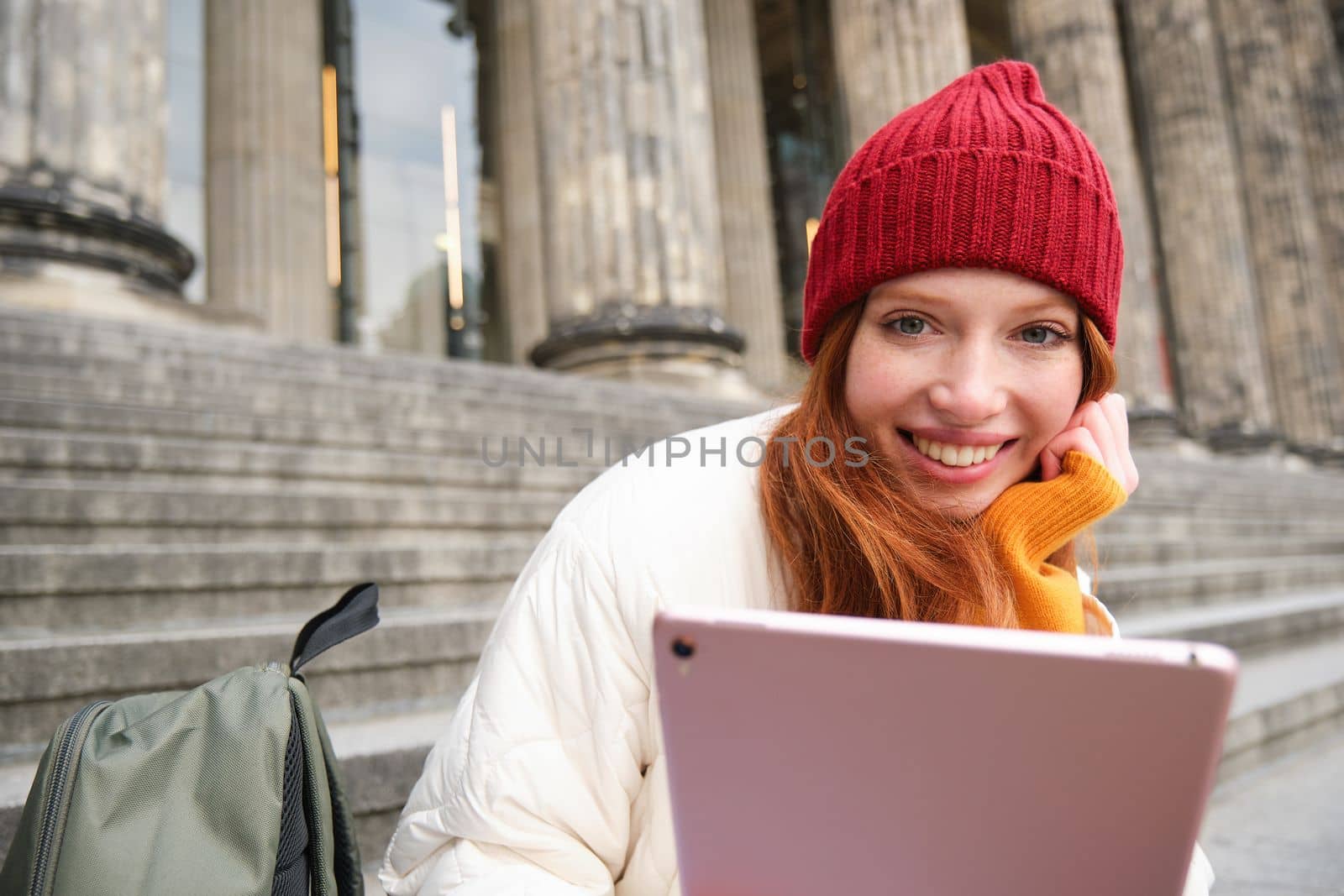Close up portrait of cute redhead european girl, sitting in red hat near municipal building on stairs, holds digital tablet, reads e-book or surfs internet.