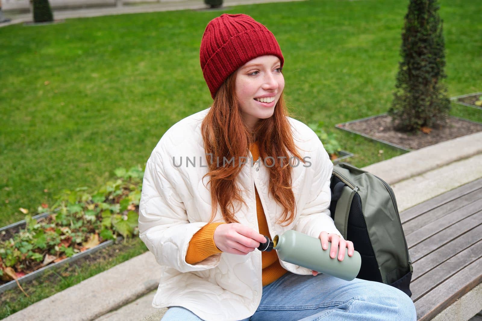 Portrait of young redhead woman in hat, sitting on bench, holding thermos, drinking hot drink from flask and smiling, resting in park.