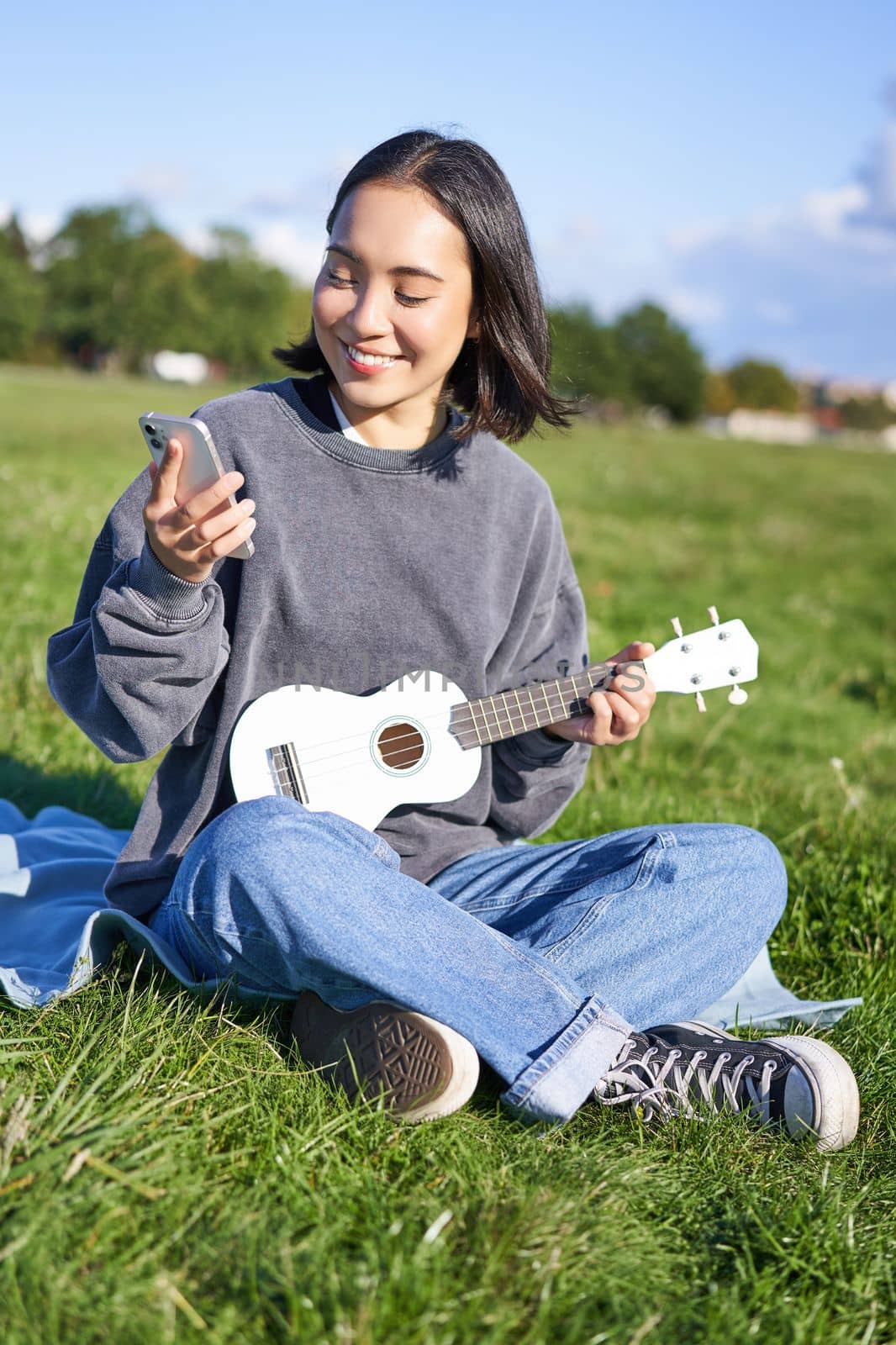 Vertical shot of smiling asian girl with smartphone, playing ukulele, reading chords or lyrics while singing, relaxing outdoors. Lifestyle and people concept by Benzoix