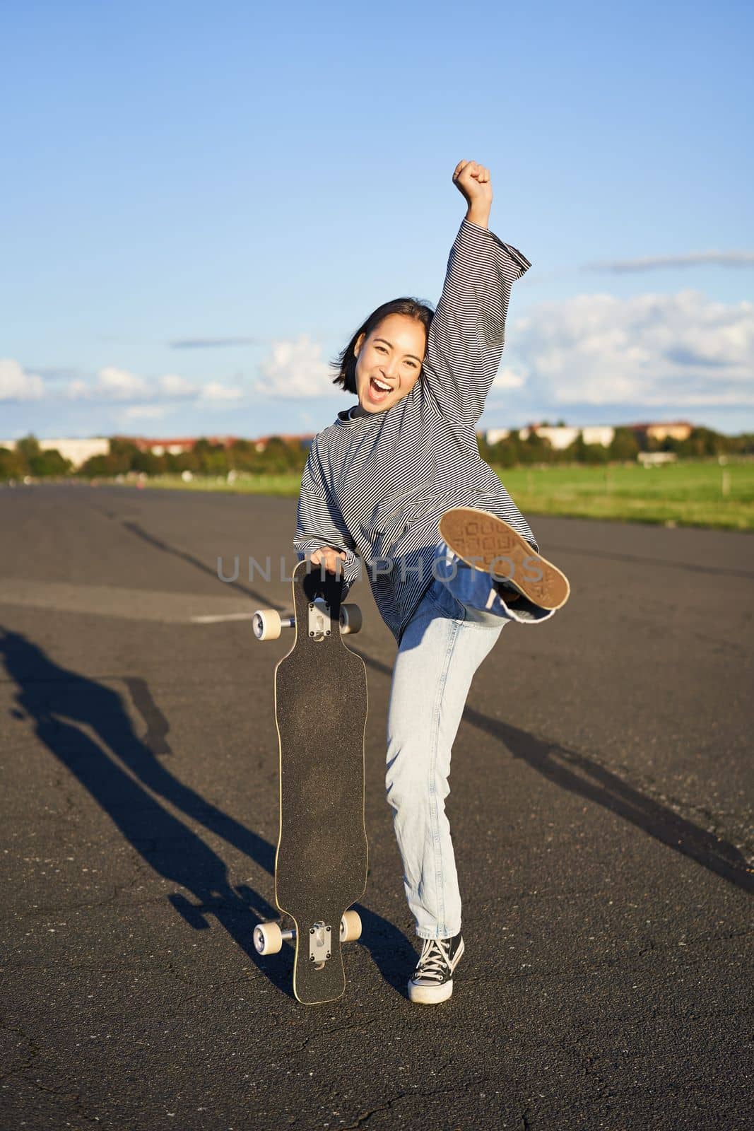 Vertical shot of asian girl feeling excited, skating on longboard, jumping and posing with skateboard, standing with cruiser on empty road, having fun outdoors.
