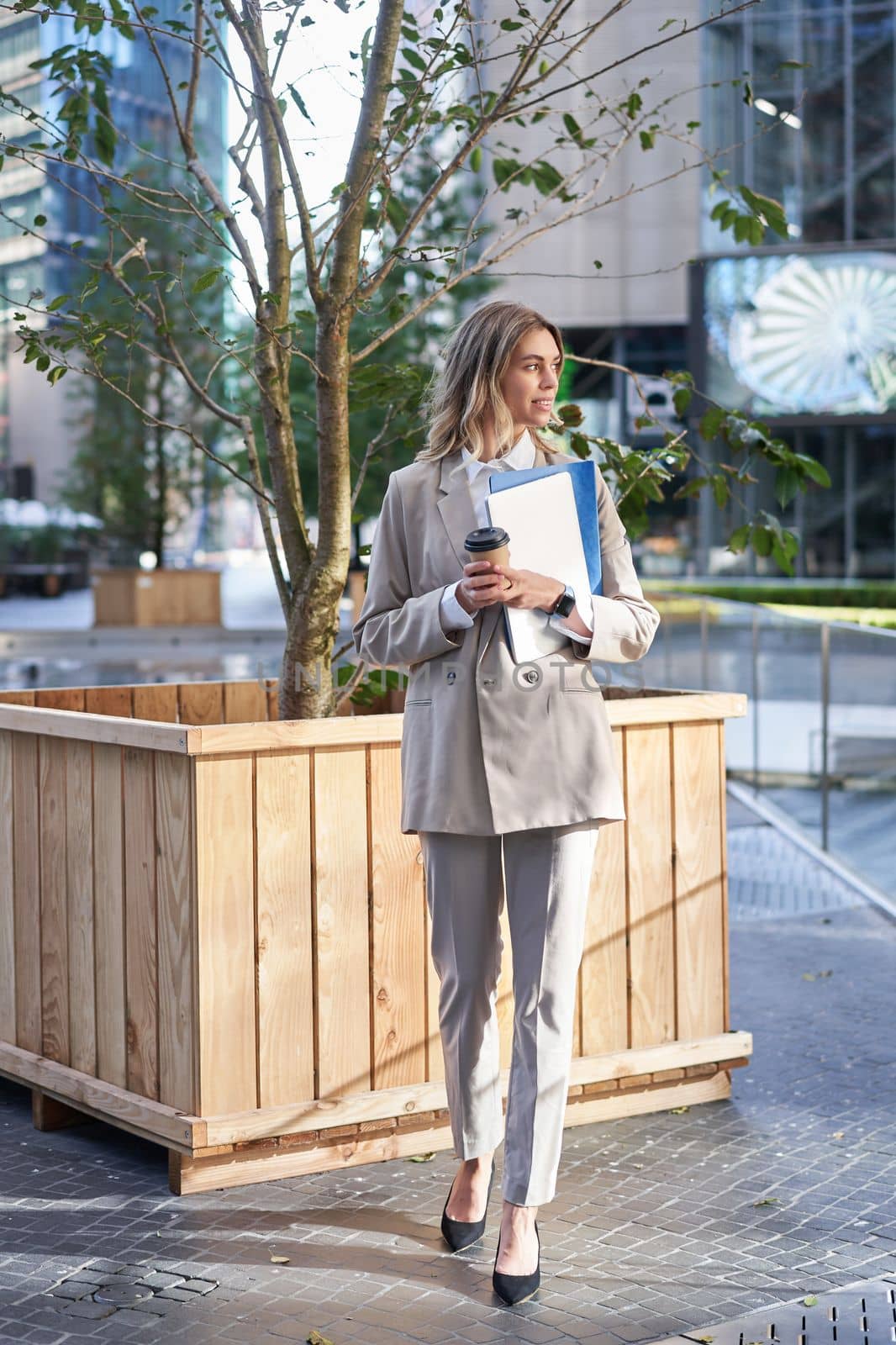 Beautiful businesswoman in suit, drinking takeaway coffee on street, waiting for someone near office building on lunch break by Benzoix