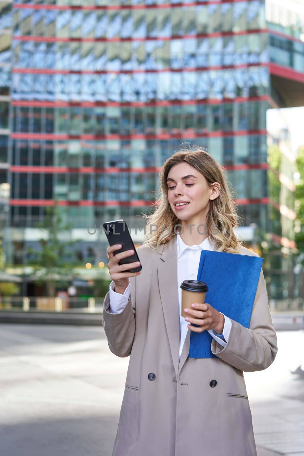 Vertical shot of businesswoman drinking coffee on street, standing in beige suit, holding documents and mobile phone by Benzoix