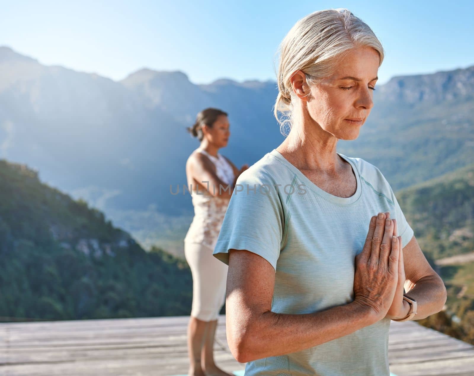 Senior woman meditating with joined hands and closed eyes breathing deeply. Class of mature people doing yoga together in nature on a sunny day.