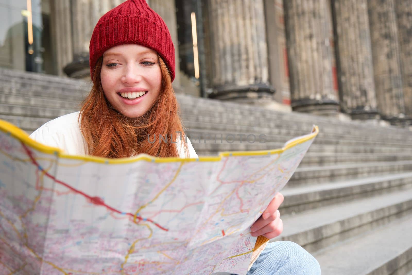 Smiling girl traveler, tourist sits on stairs near museum and holds paper map, explores city during sightseeing adventure trip by Benzoix