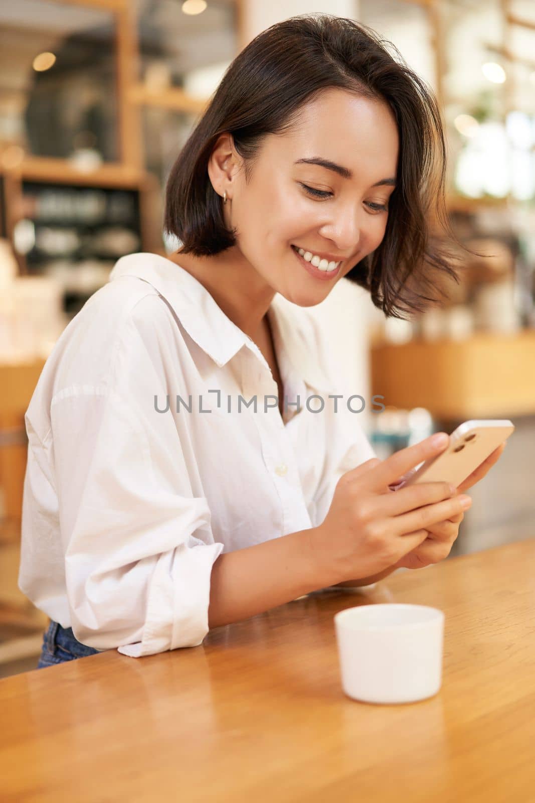 Portrait of young brunette woman, sitting with coffee and using smartphone in a cafe, chatting on mobile phone.