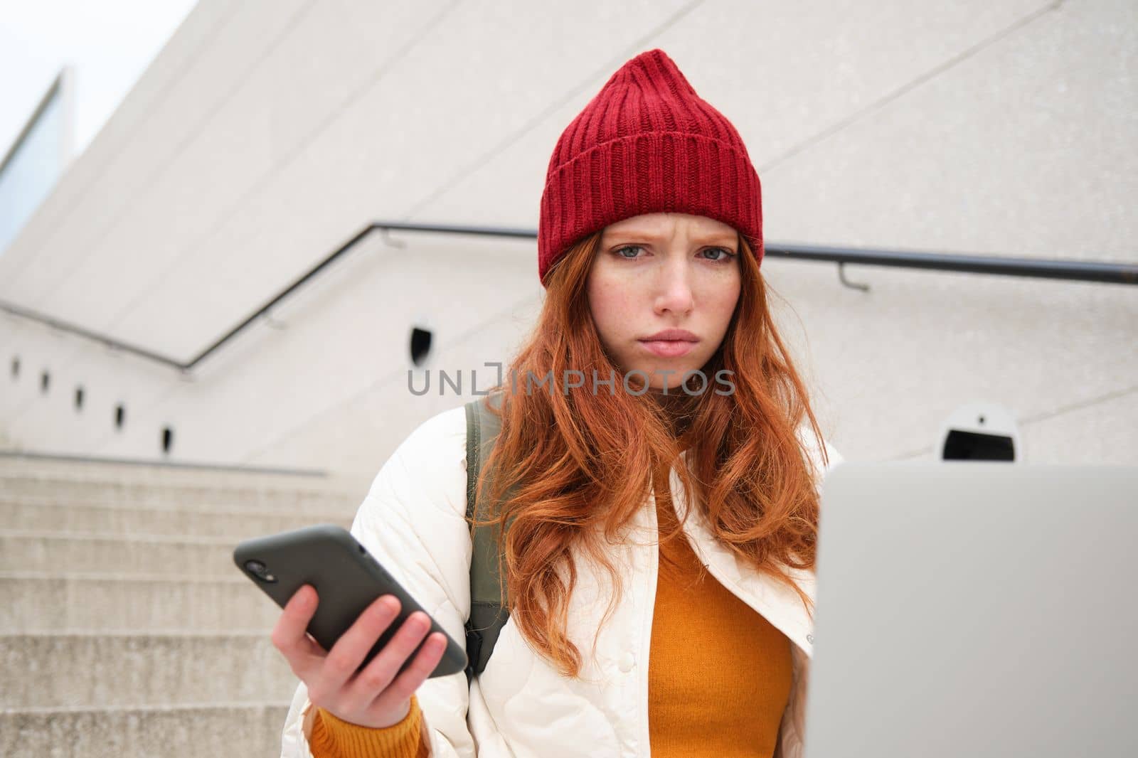 Portrait of redhead woman sits on stairs, uses laptop and holds smartphone, looks confused and upset at computer screen, tries to connect to wifi public internet by Benzoix