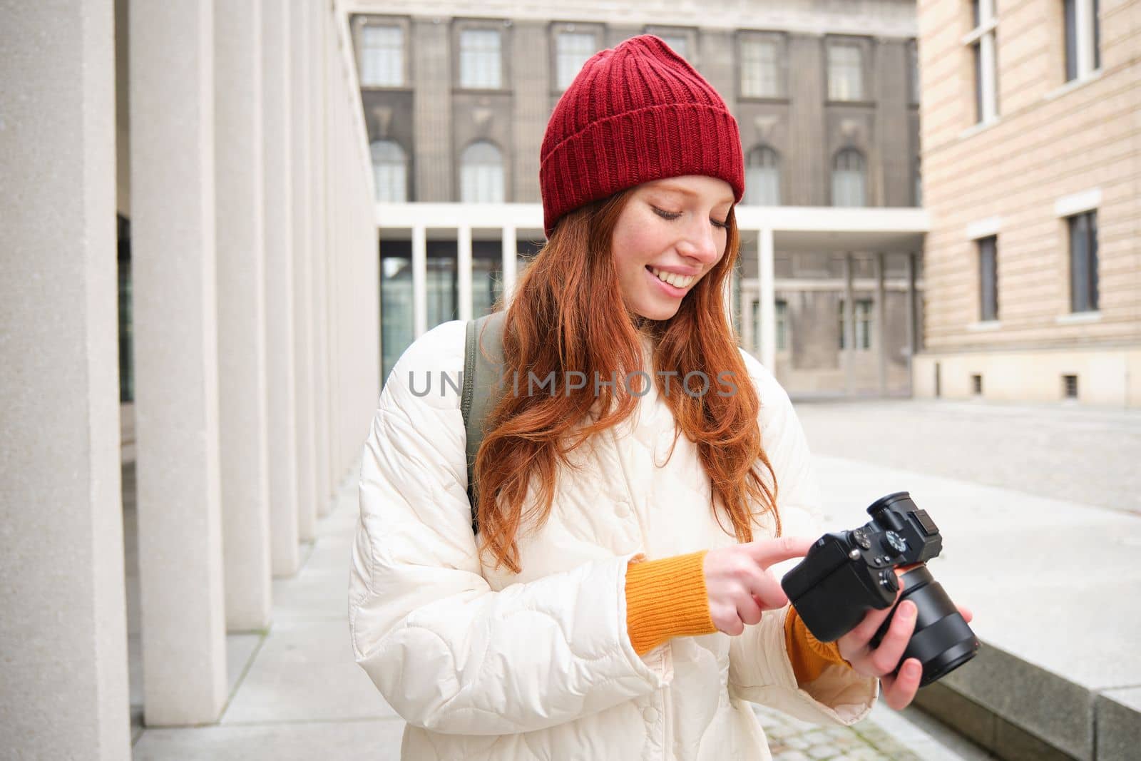 Smiling redhead girl photographer, taking pictures in city, makes photos outdoors on professional camera. Young talent and hobby concept