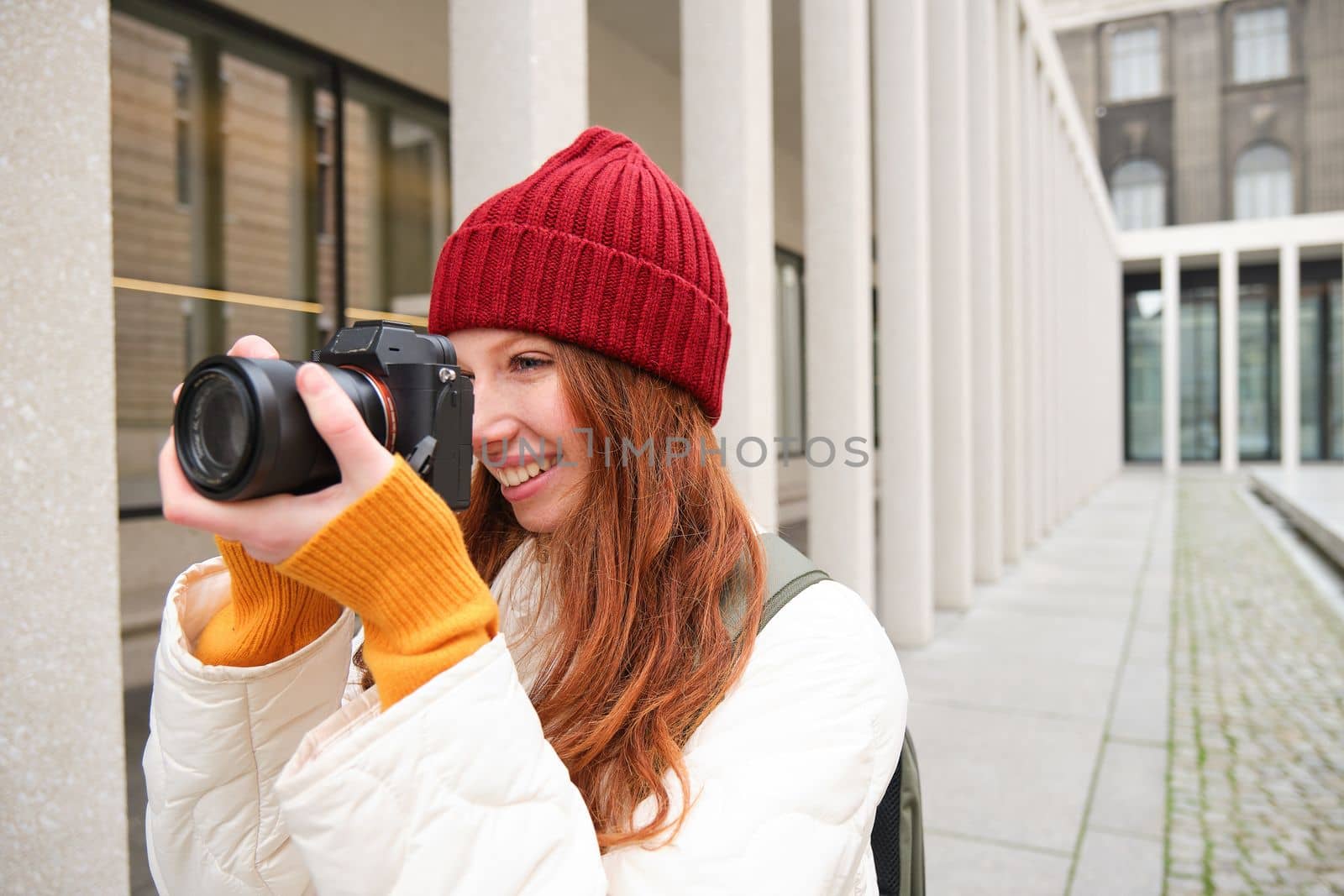 Smiling redhead girl photographer, taking pictures in city, makes photos outdoors on professional camera by Benzoix
