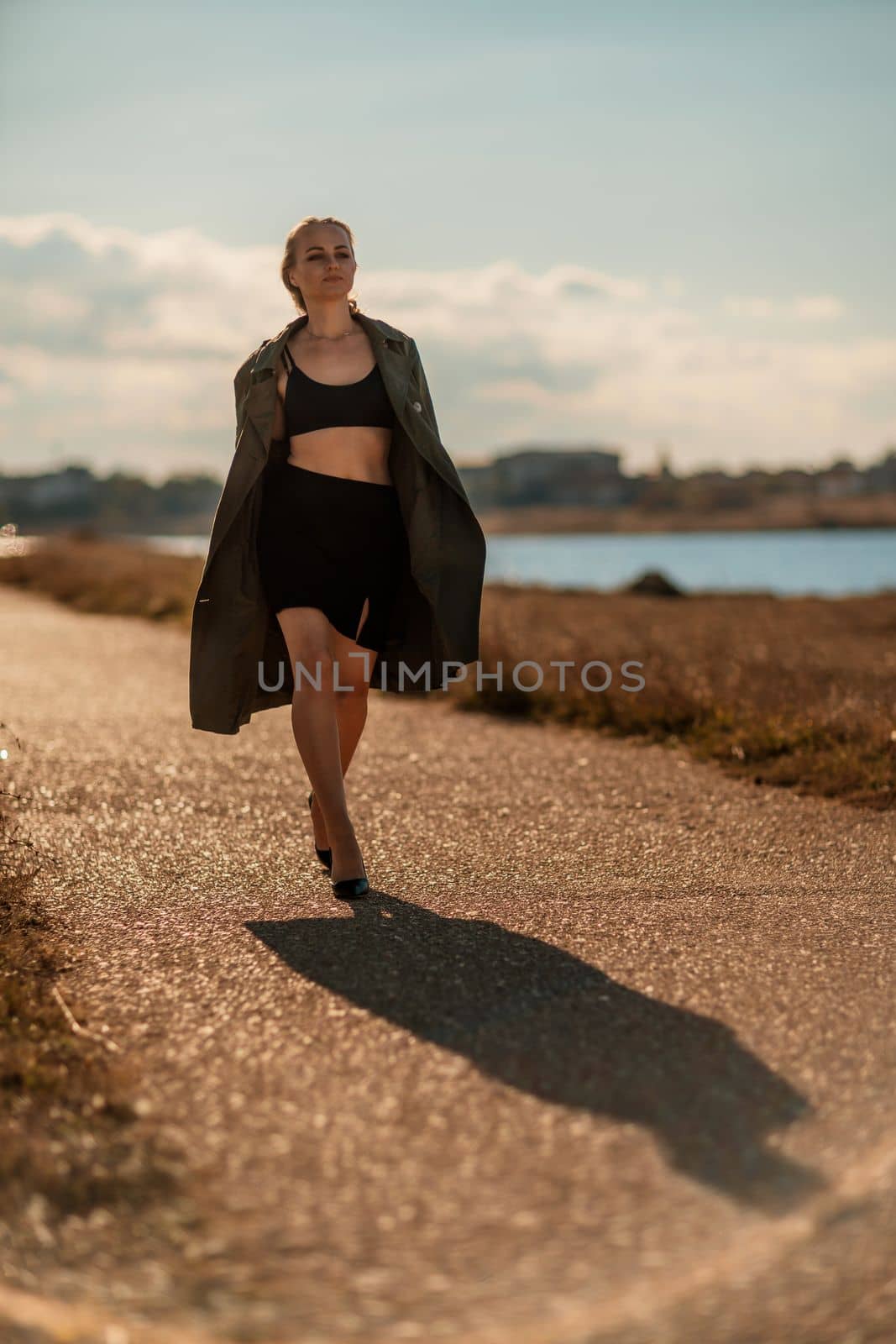 Portrait blonde sea cape. A calm young blonde in an unbuttoned khaki raincoat walks along the seashore, under a raincoat a black skirt and top.