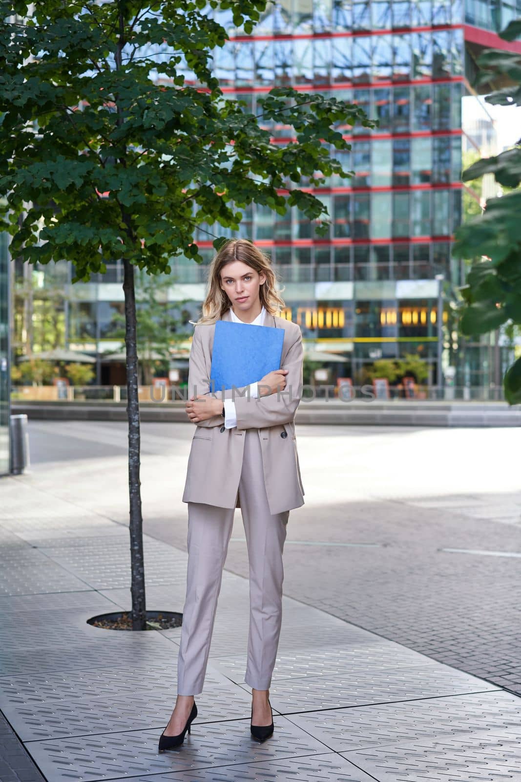 Vertical shot of young businesswoman in beige suit, holding documents in hands, looking confident at camera, standing outdoors in city.