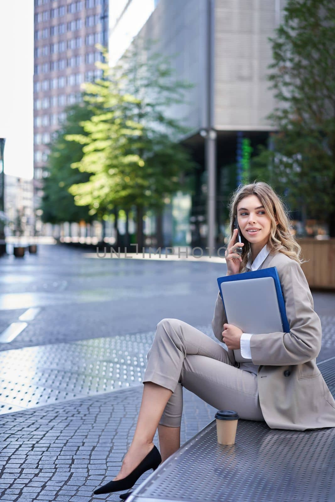 Vertical shot of young woman sitting near office building, holding laptop and folders with work documents, calling someone on mobile phone.