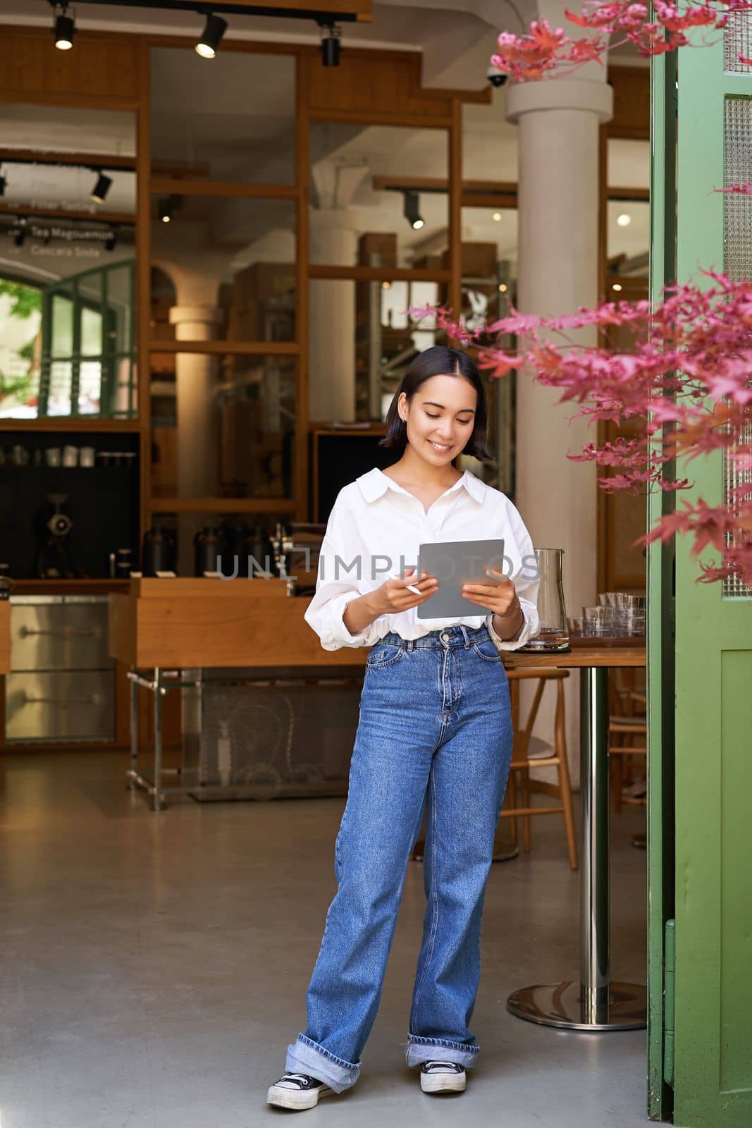 Vertical shot of smiling asian businesswoman, waitress standing in front of cafe entrance, inviting guests in restaurant with tablet by Benzoix