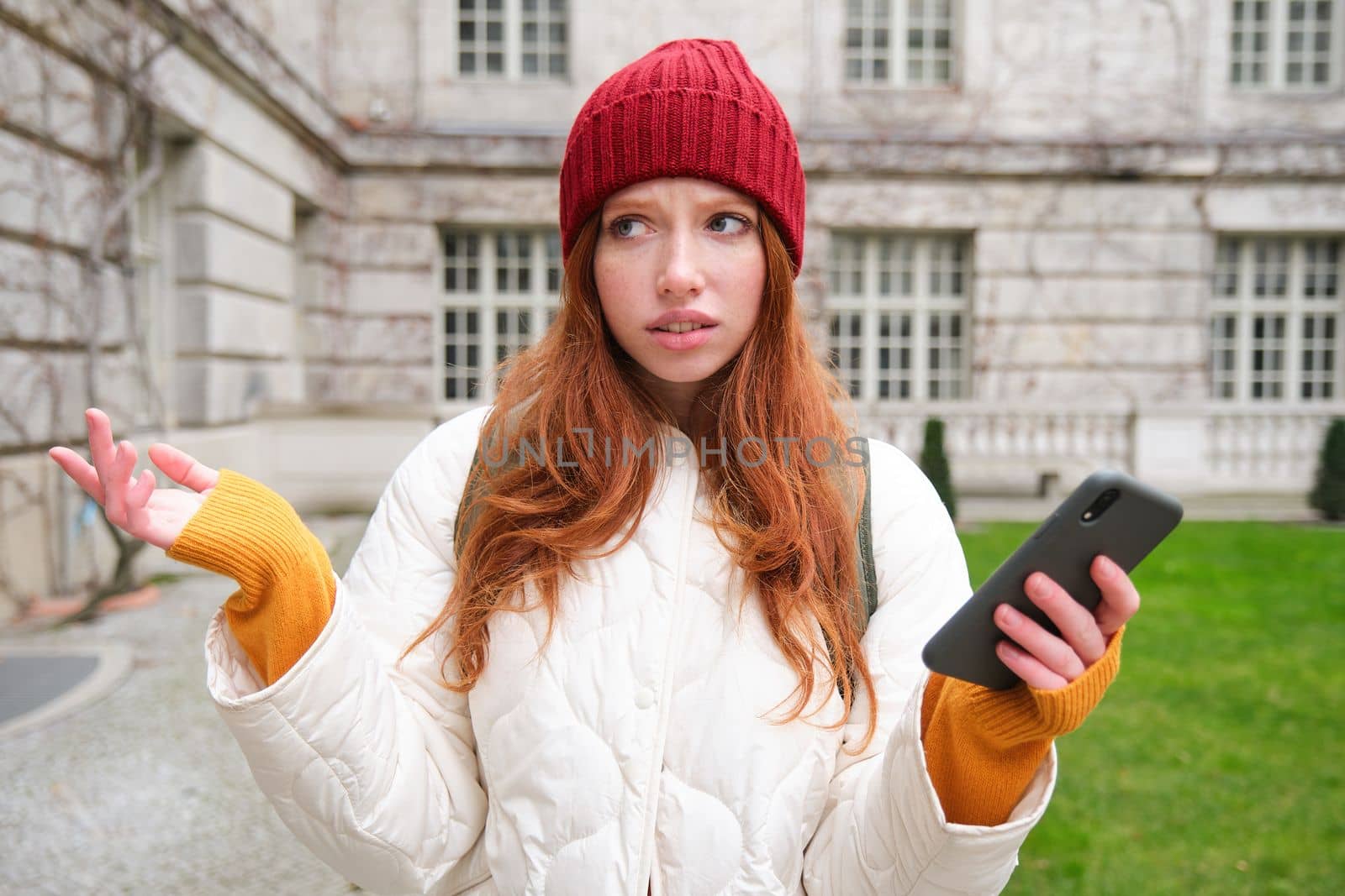 Portrait of confused girl tourist with smartphone, shrugs puzzled, dont know where to go, holds mobile phone with map application by Benzoix