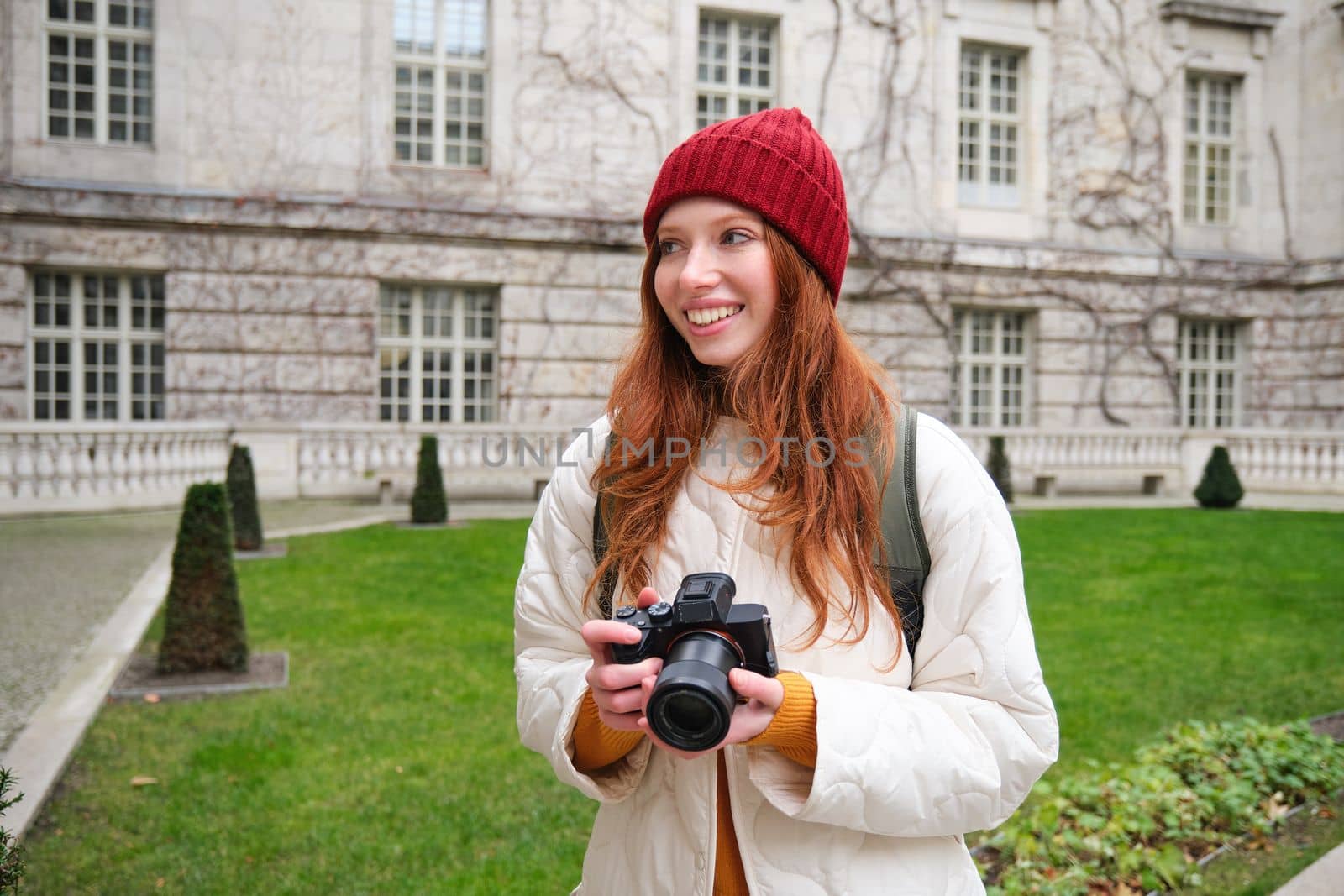 Redhead girl photographer takes photos on professional camera outdoors, captures streetstyle shots, looks excited while taking pictures.