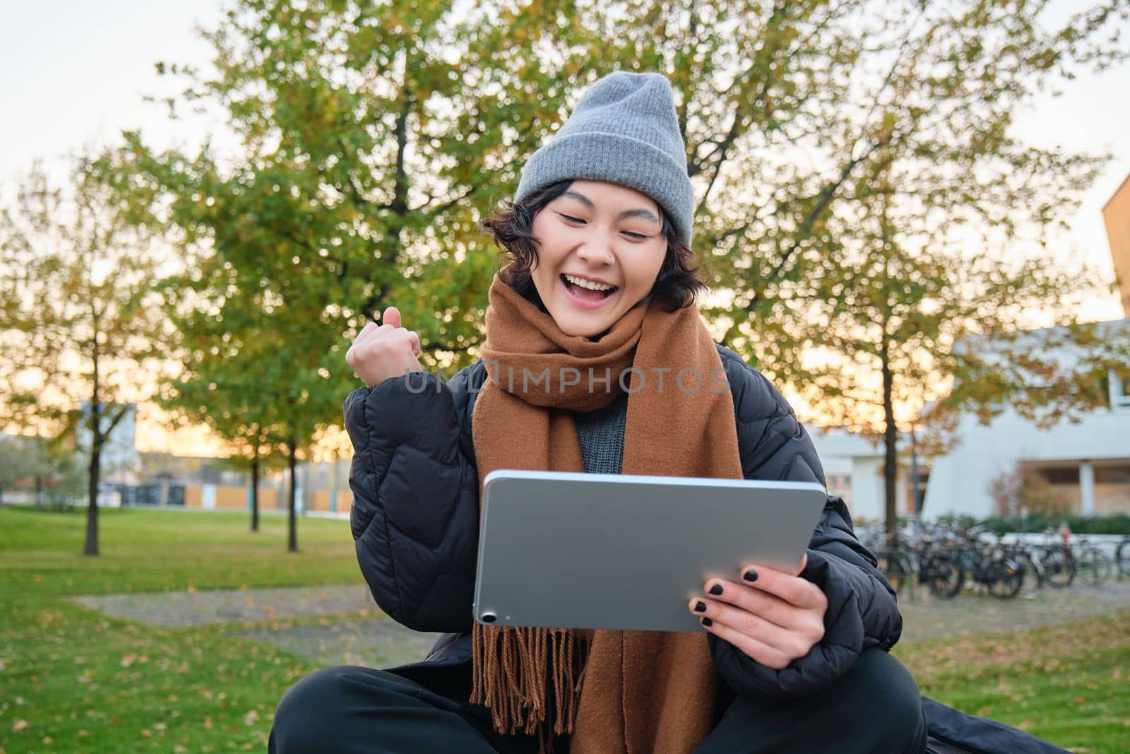 Portrait of happy asian girl sits on bench, looks at digital tablet screen and cheers, triumphs, wins something, celebrates good news, relaxes in park outdoors.