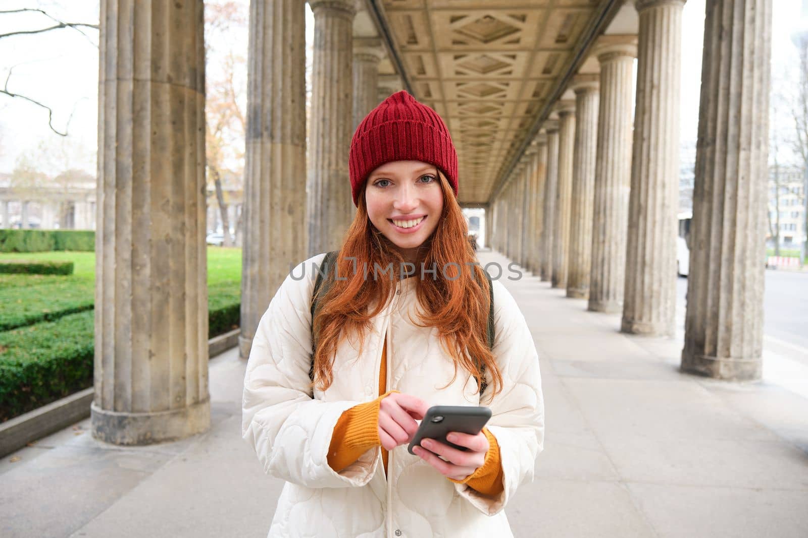 Mobile broadband and people. Smiling redhead 20s girl with backpack, uses smartphone on street, holds mobile phone and looks at application.
