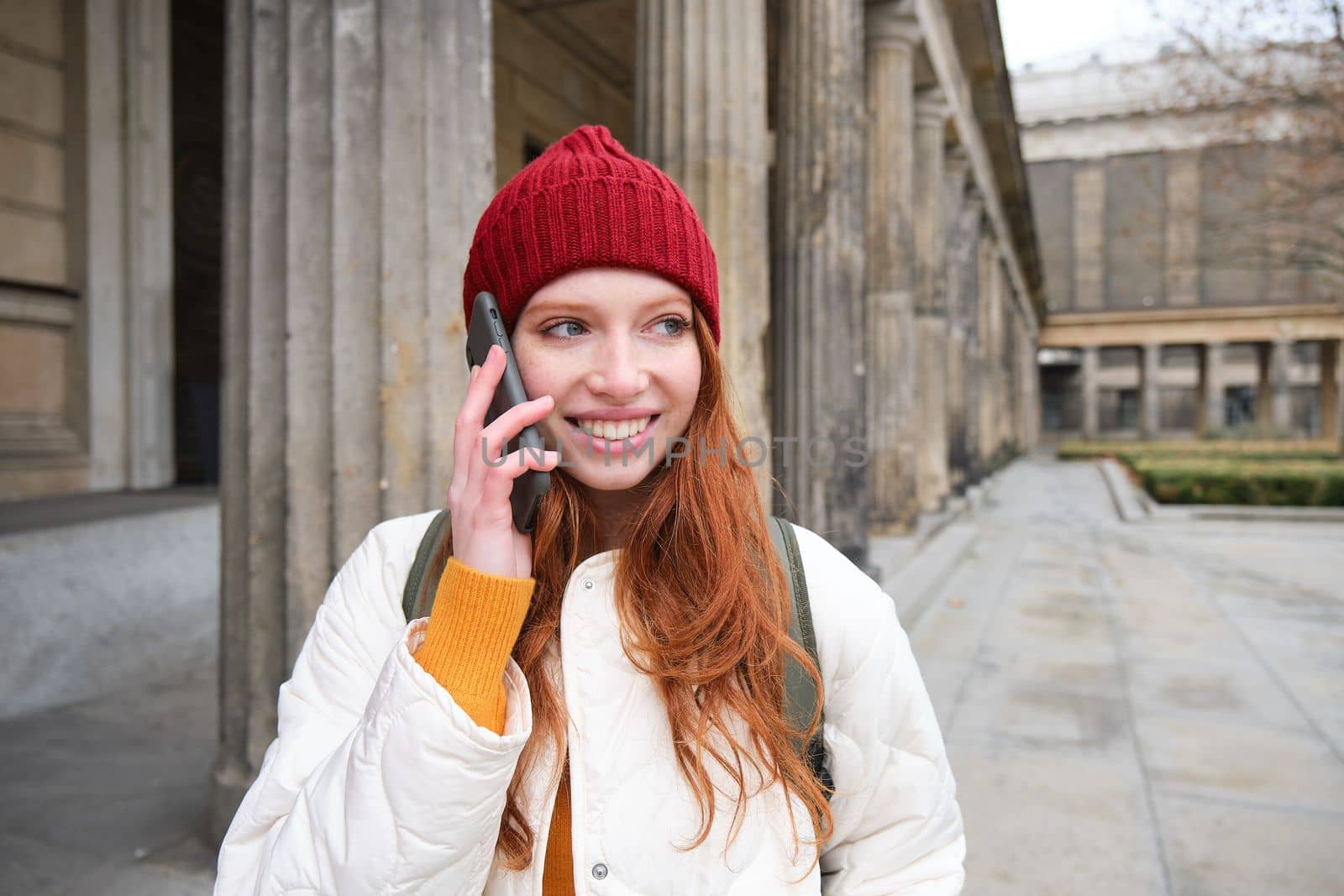 Smiling redhead female tourist talks on mobile phone and walks around city. Happy student in red hat calls friend, stands on street and uses smartphone.