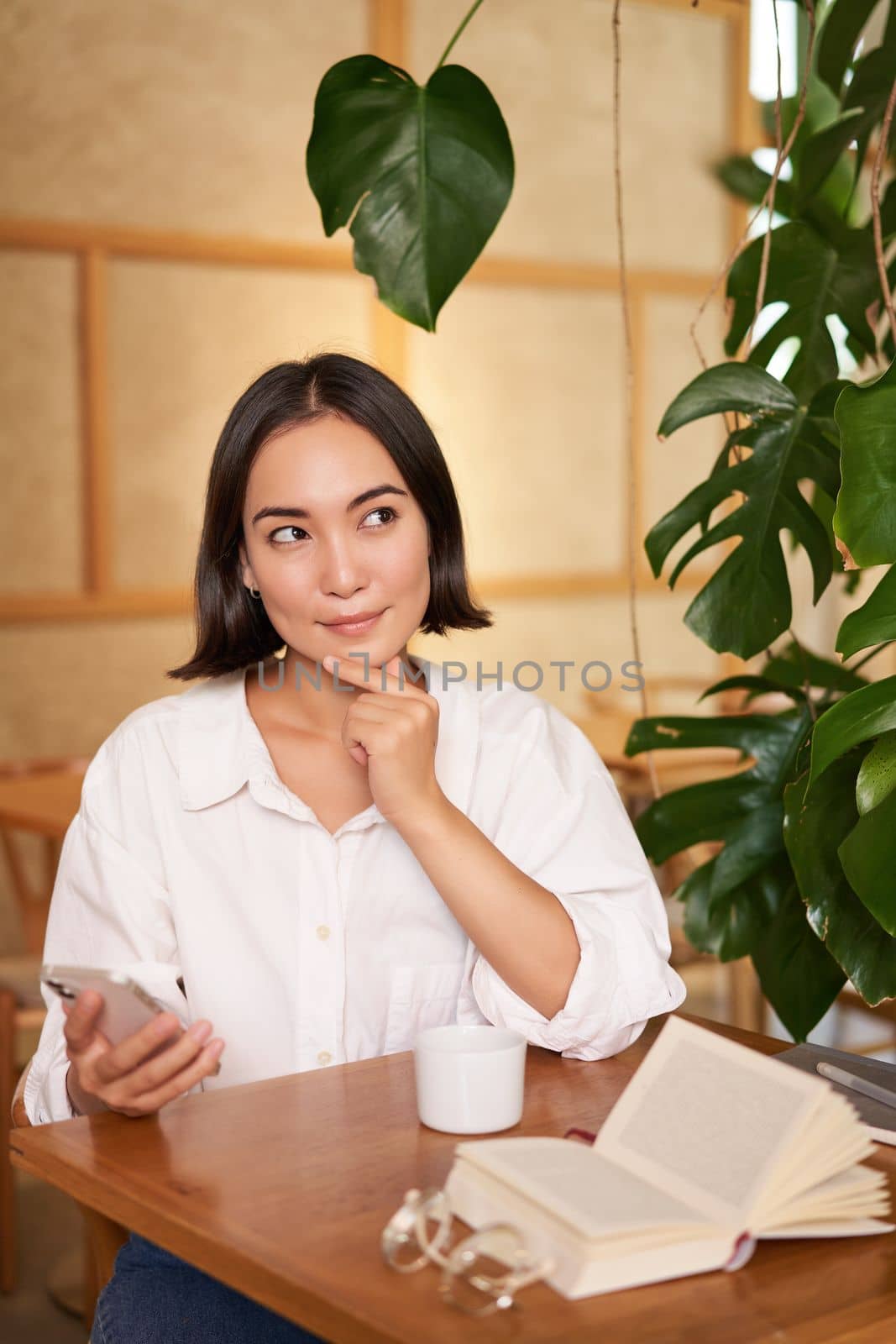 Beautiful young woman, 25 years old, thinking, holding smartphone and looking thoughtful, sitting in cafe, deciding smth.