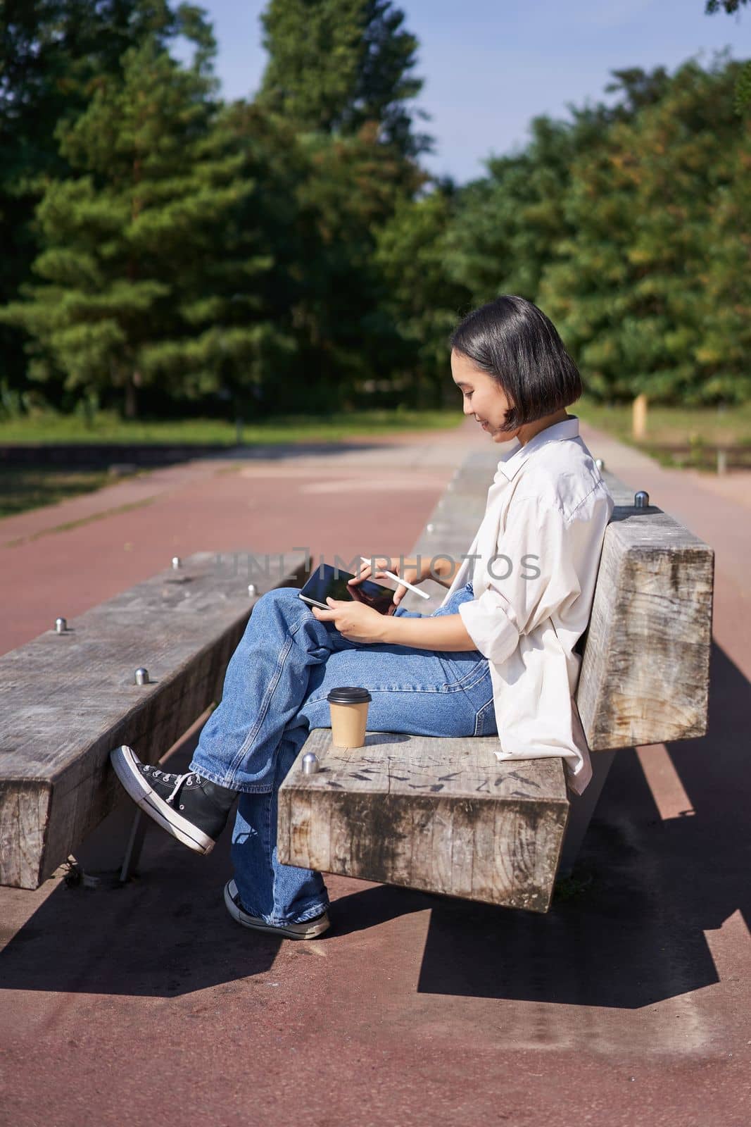 Asian girl, drawing in park, sitting on street on sunny day with digital graphic tablet and pen, smiling happily.