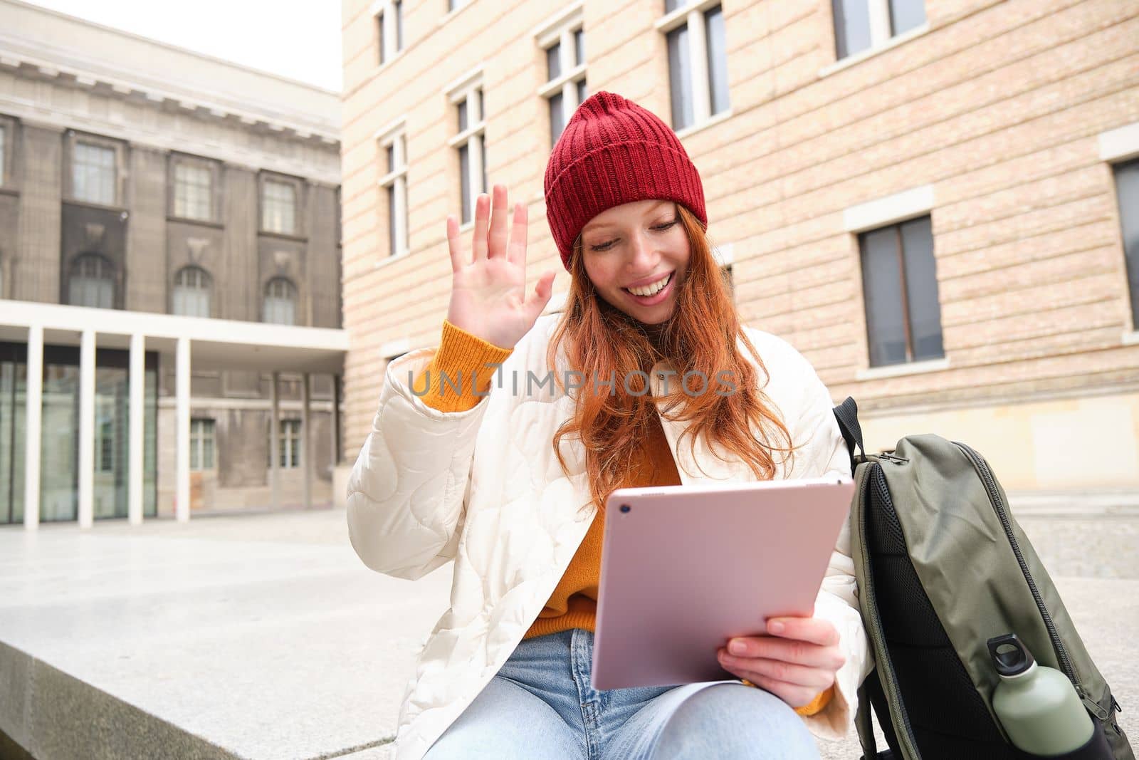 Happy redhead girl, student sits with backpack and digital tablet on street, says hello and waves at gadget camera, connects to public wifi to video chat, has online conversation by Benzoix
