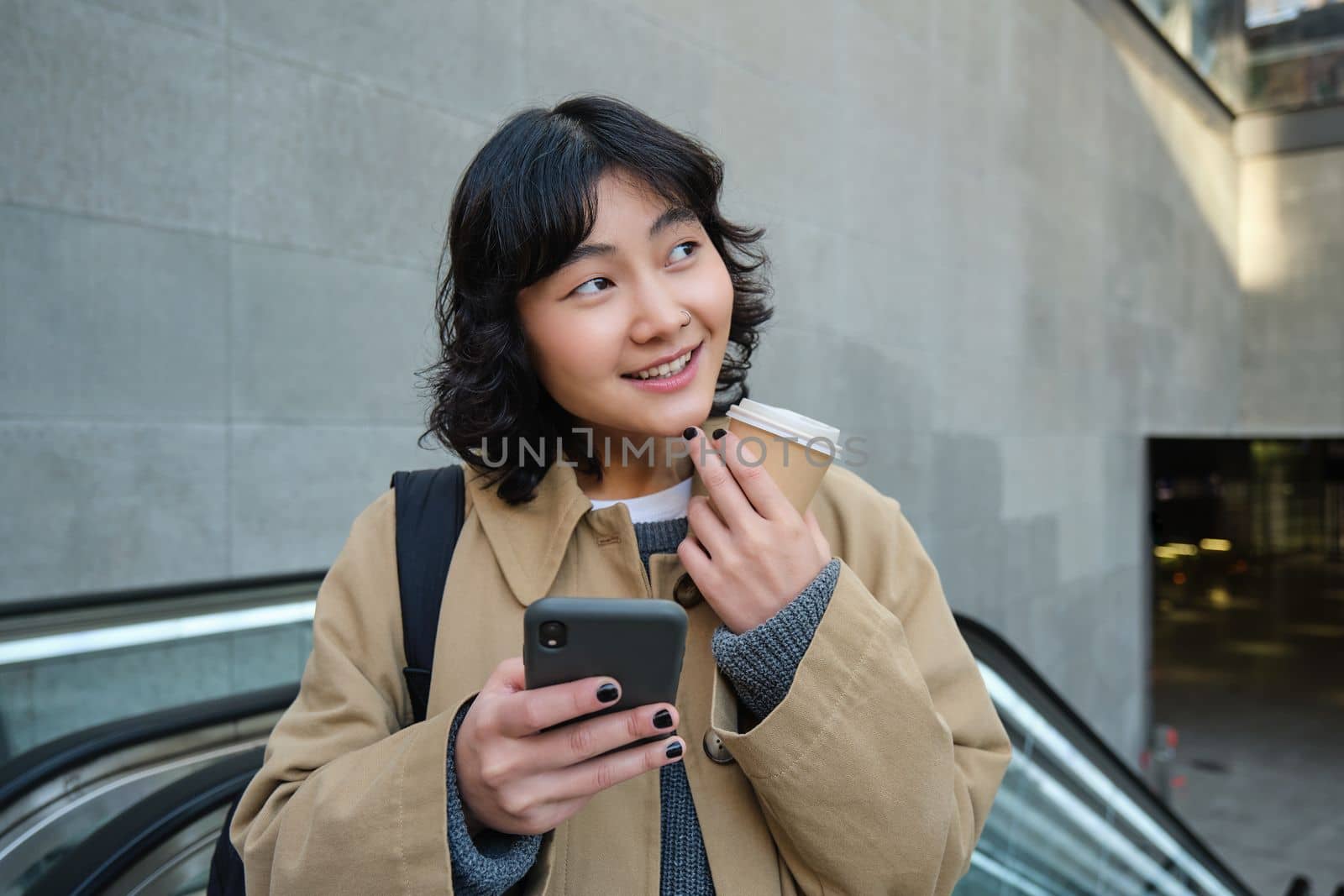 Beautiful happy asian girl, drinks coffee to go, using mobile phone while standing on escalator, walking in city centre and smiling.