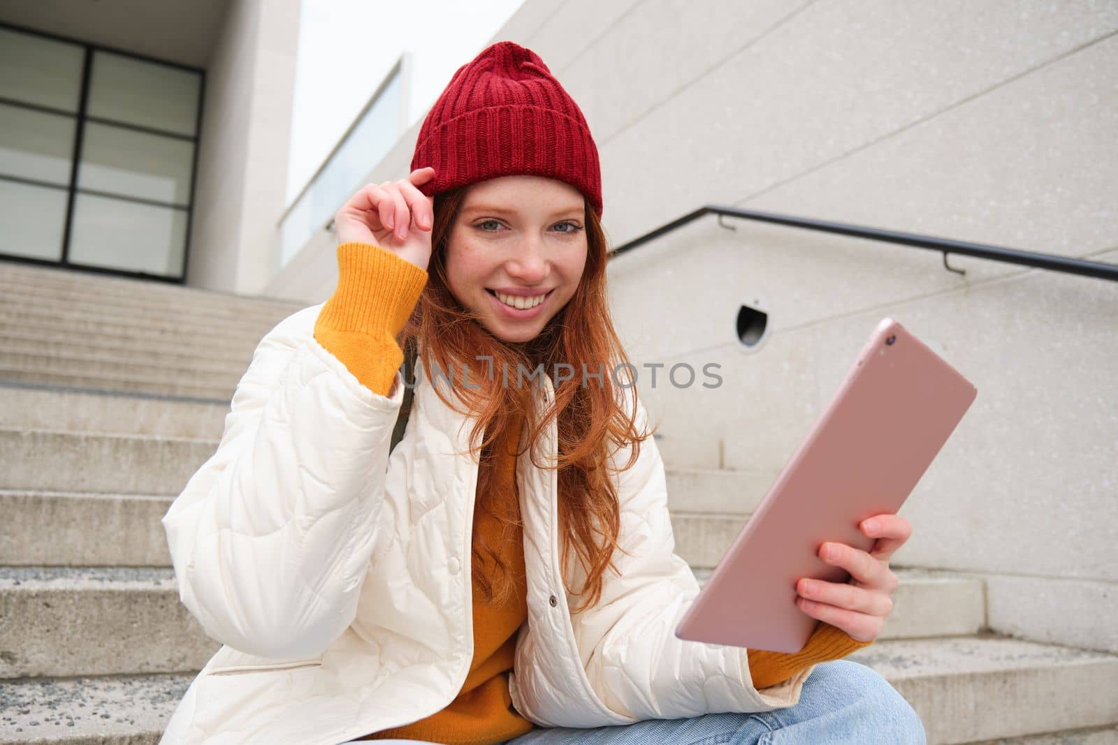 Young stylish girl, redhead female students sits on stairs outdoors with digital tablet, reads, uses social media app on gadget, plays games while waits on street by Benzoix