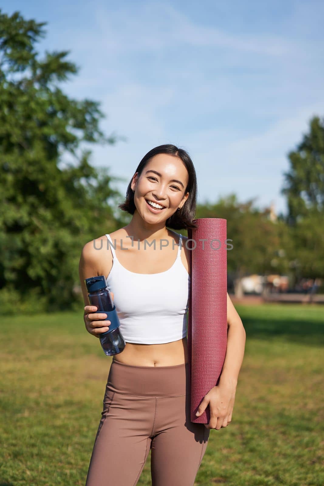Portrait of young slim and healthy korean girl doing workout in park, standing with water bottle and rubber mat for execises on green lawn, smiling happily.