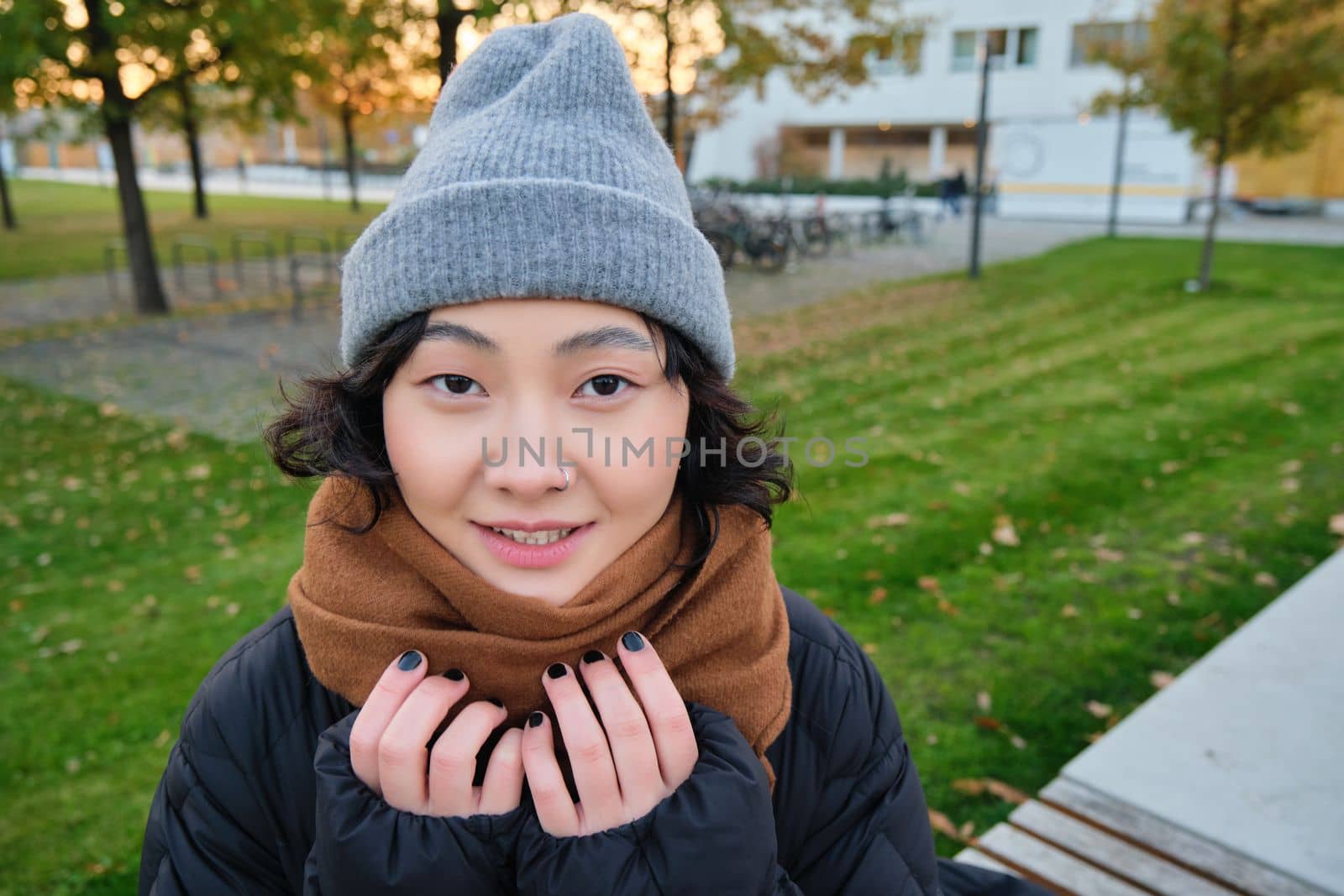 Portrait of cute asian girl in hat and scarf, walks around town in chilly spring weather, smiles and looks happy, sits in park near green grass.