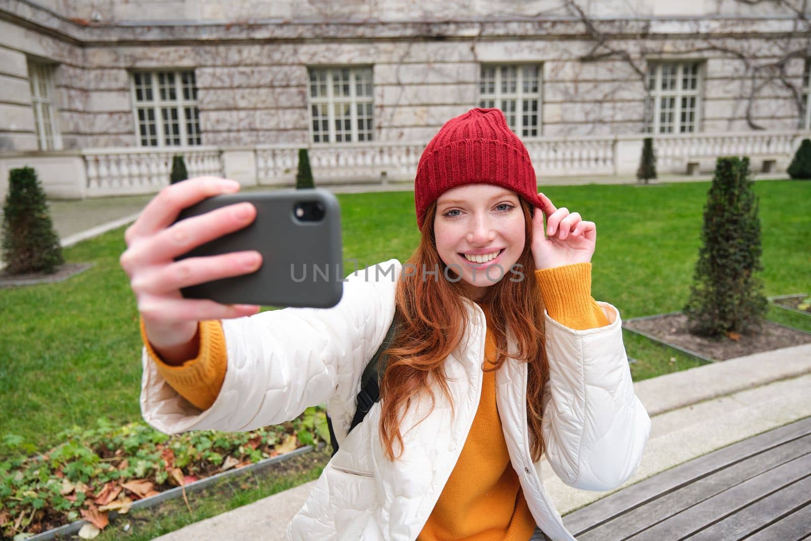 Beautiful smiling ginger girl in hat, posing for photo on mobile phone, takes selfie in park with cute face expression by Benzoix