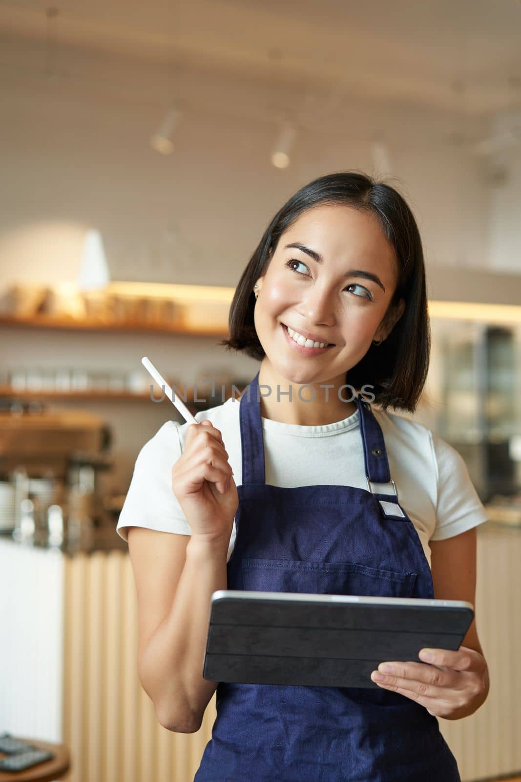 Vertical shot of smiling girl waitress, barista in coffee shop, wears blue apron uniform, takes order with tablet, stands in cafe.