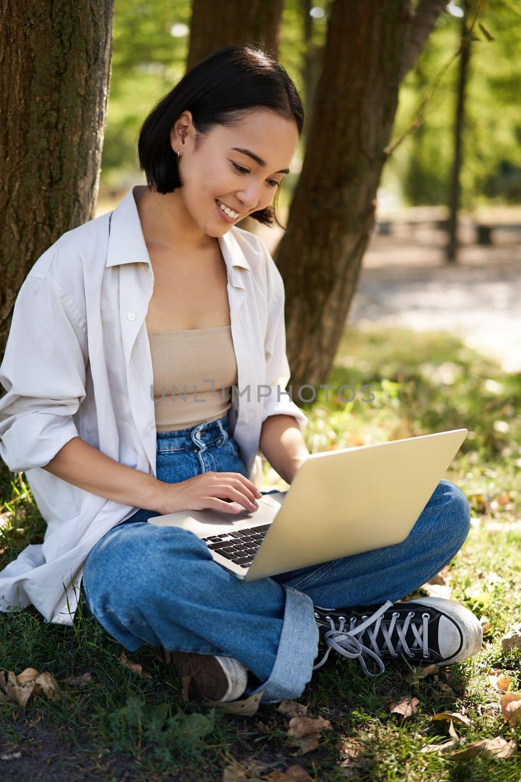 Beautiful asian girl sitting in park with laptop, working on remote, typing on keyboard, smiling at camera, resting beside tree.