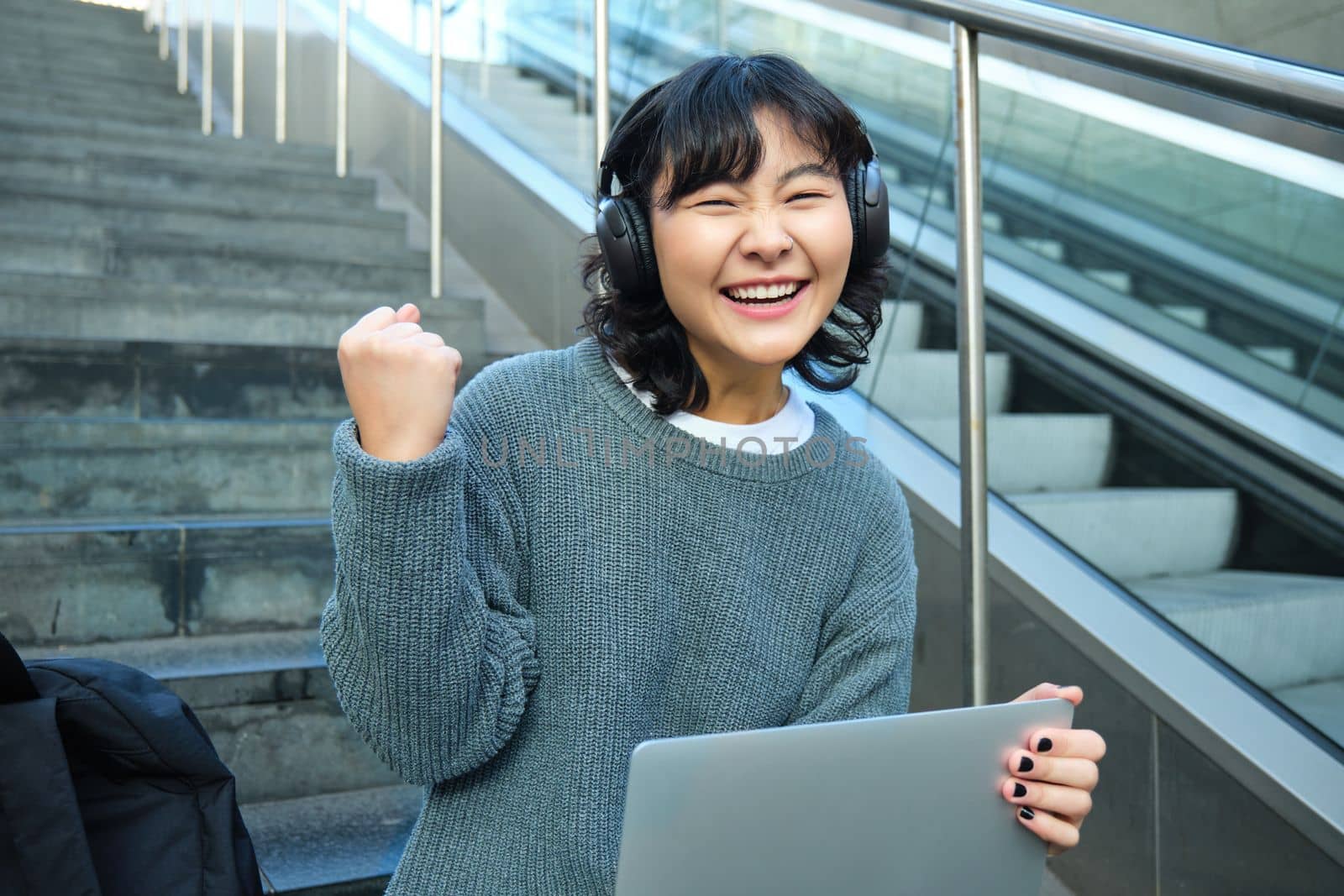 Cheerful korean girl in headphones, sits on stairs with laptop, celebrates, say yes, triumphs, receives good news, success on working project, works remotely on street.