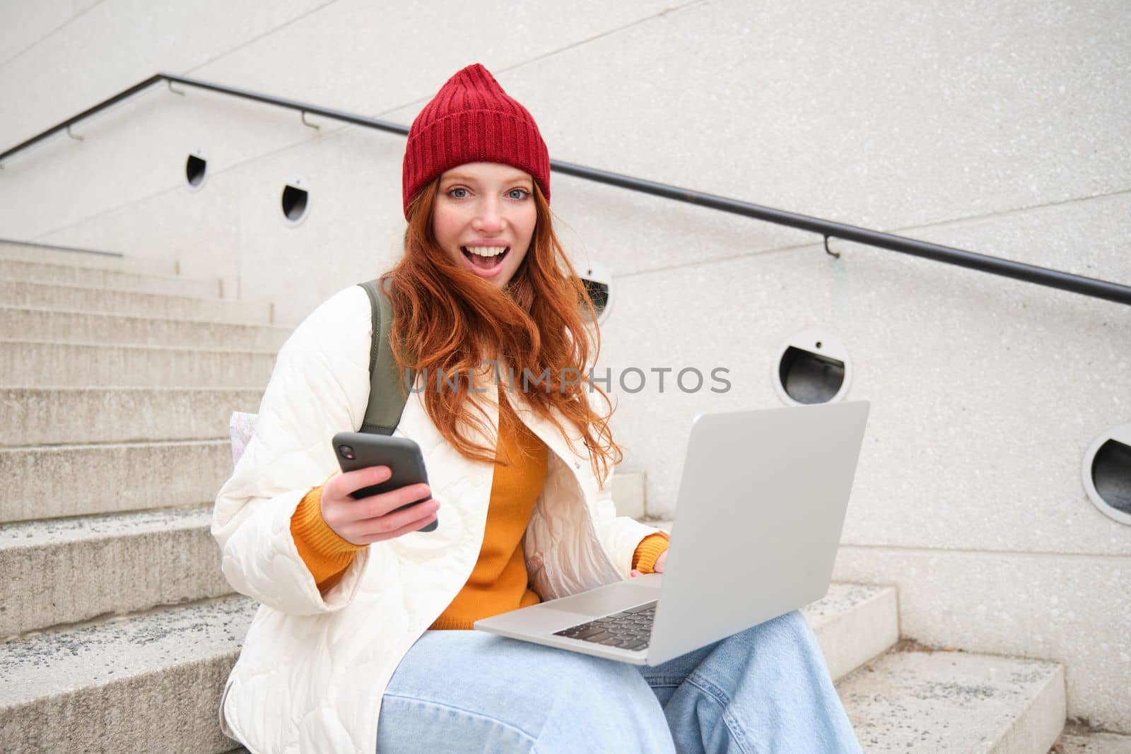 Young tourist, girl booking hotel room on laptop, enters confirmation code on smartphone, sitting with computer and phone on stairs outdoors.