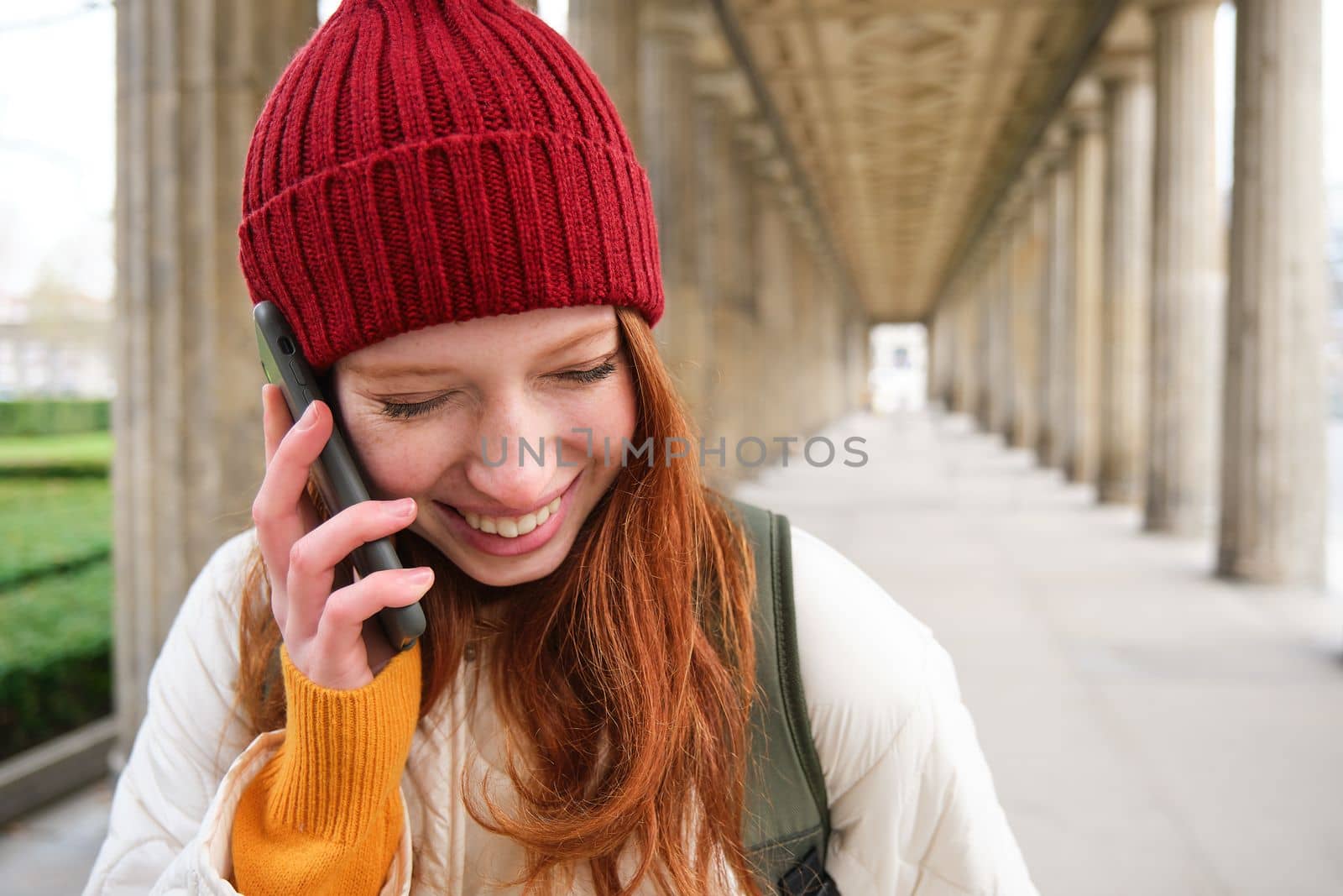 Portrait of redhead european girl in red hat, makes a phone call, walks in city and talks to friend on smartphone. Copy space