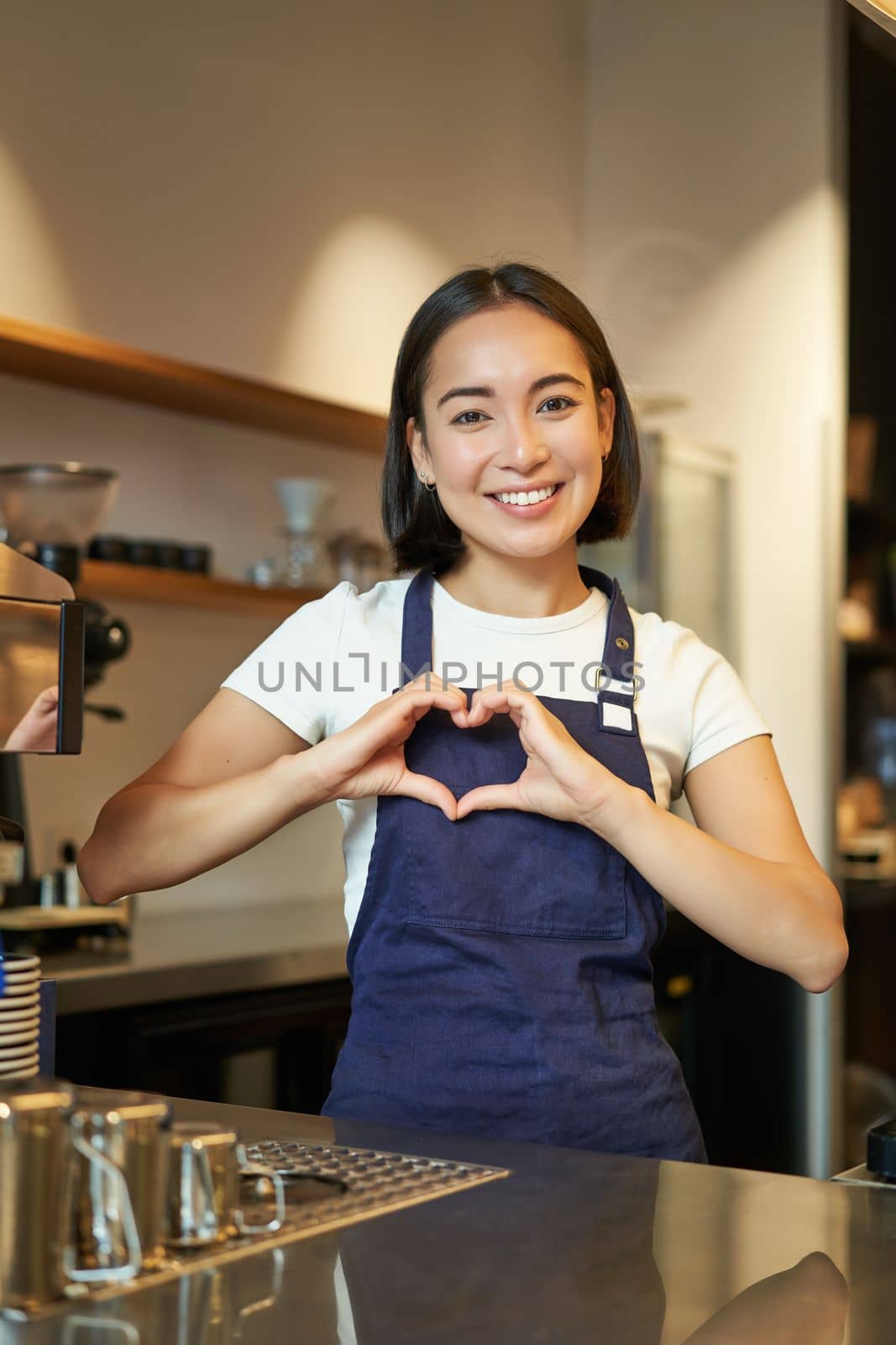 Smiling asian girl barista, shows heart sign, loves making coffee an serving clients, standing in uniform behind counter, work in cafe.