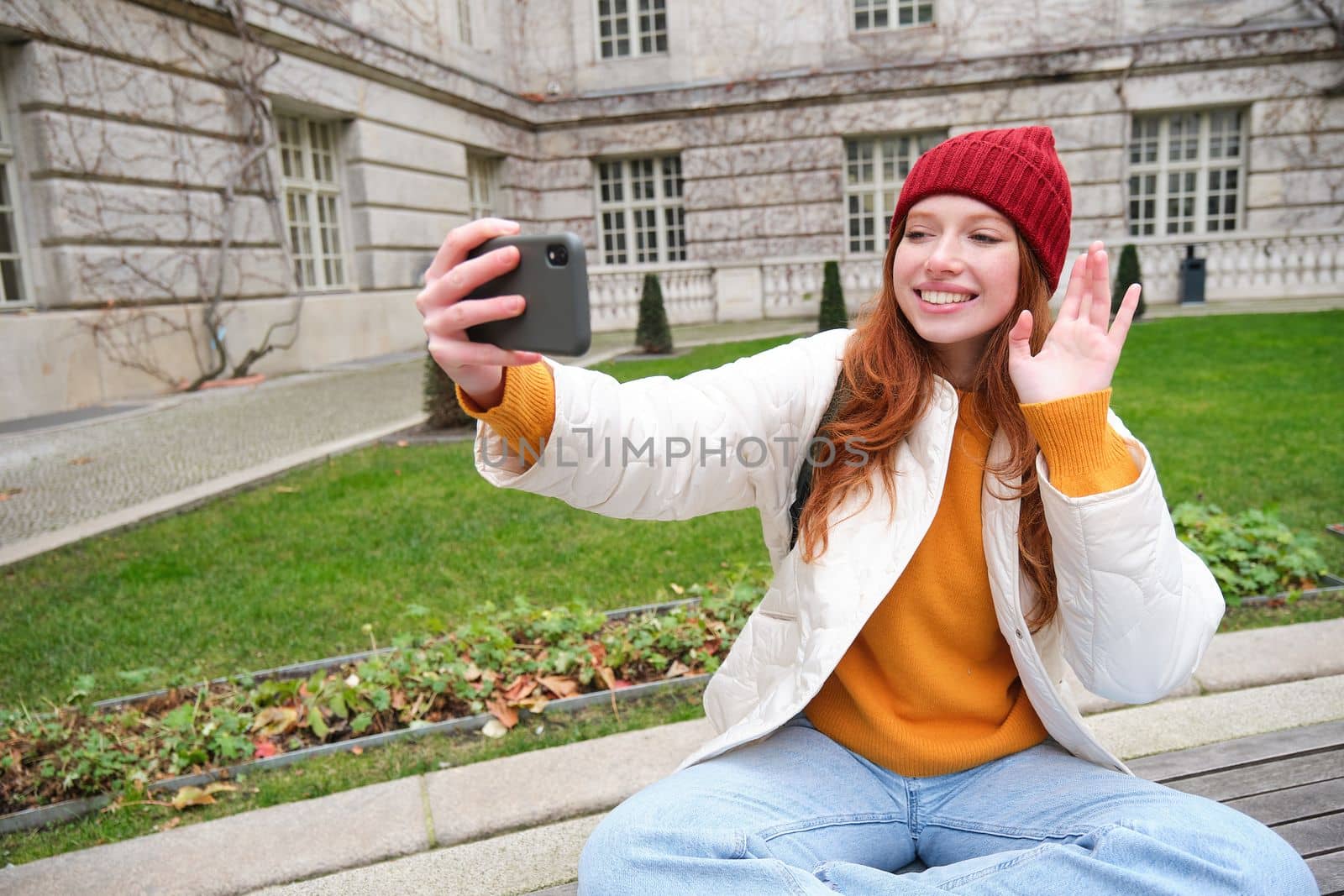 Young teen redhead girl sits on bench in park and takes selfie, makes a photo of herself with smartphone app, records vlog.