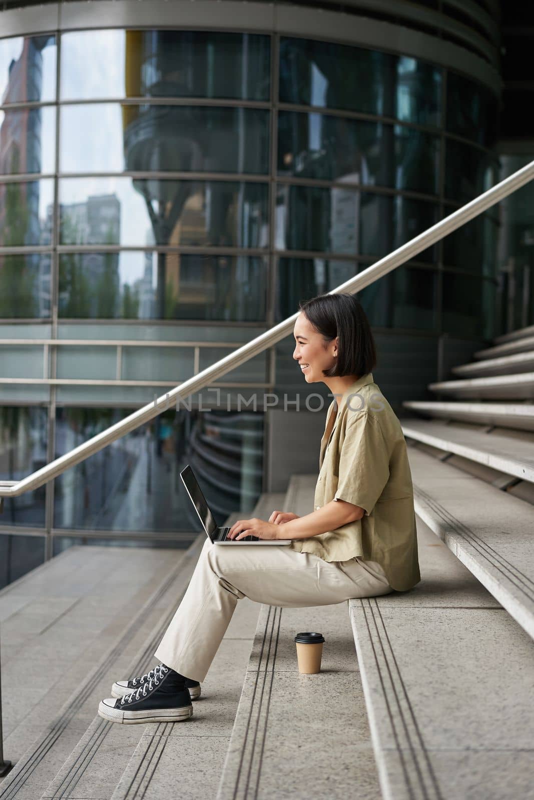 Profile portrait of young asian woman with laptop, girl student sits on stairs outside building and types on computer, drinks takeaway coffee.