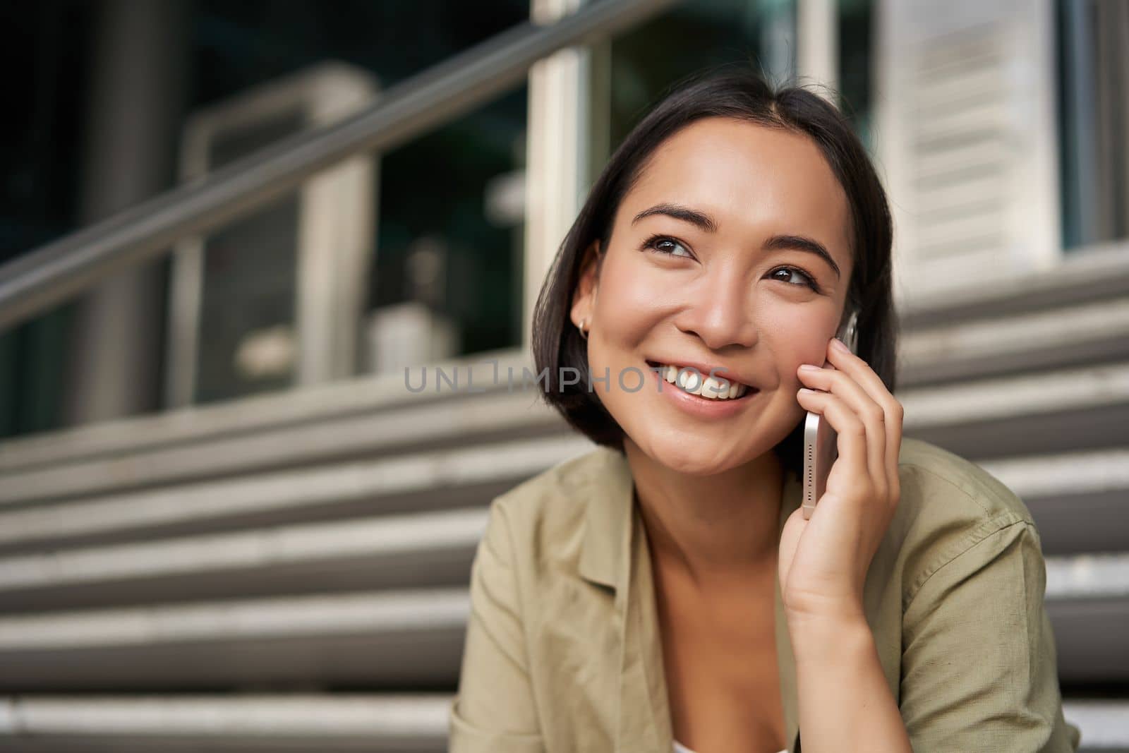 Portrait of asian girl smiles while talks on mobile phone. Young woman calling a friend, sitting on stairs. Technology concept