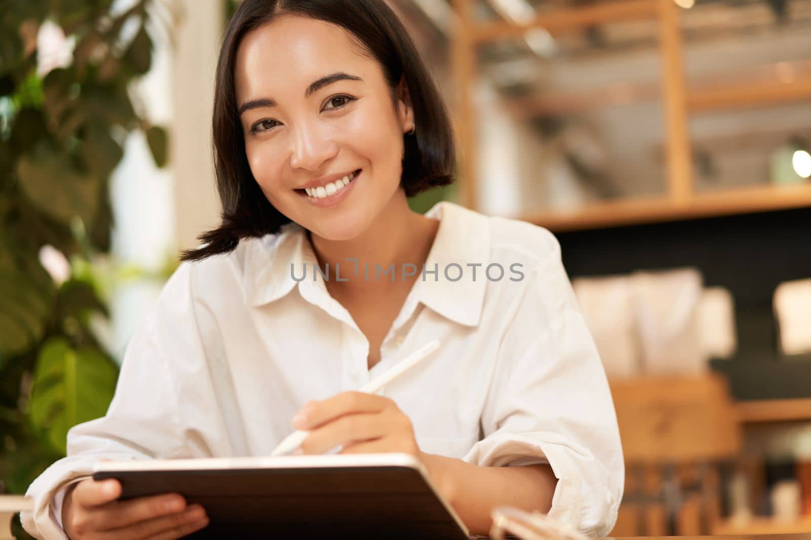 Portrait of asian girl drawing on digital tablet, scatching and doodling with graphic pen, sitting in cafe and smiling.