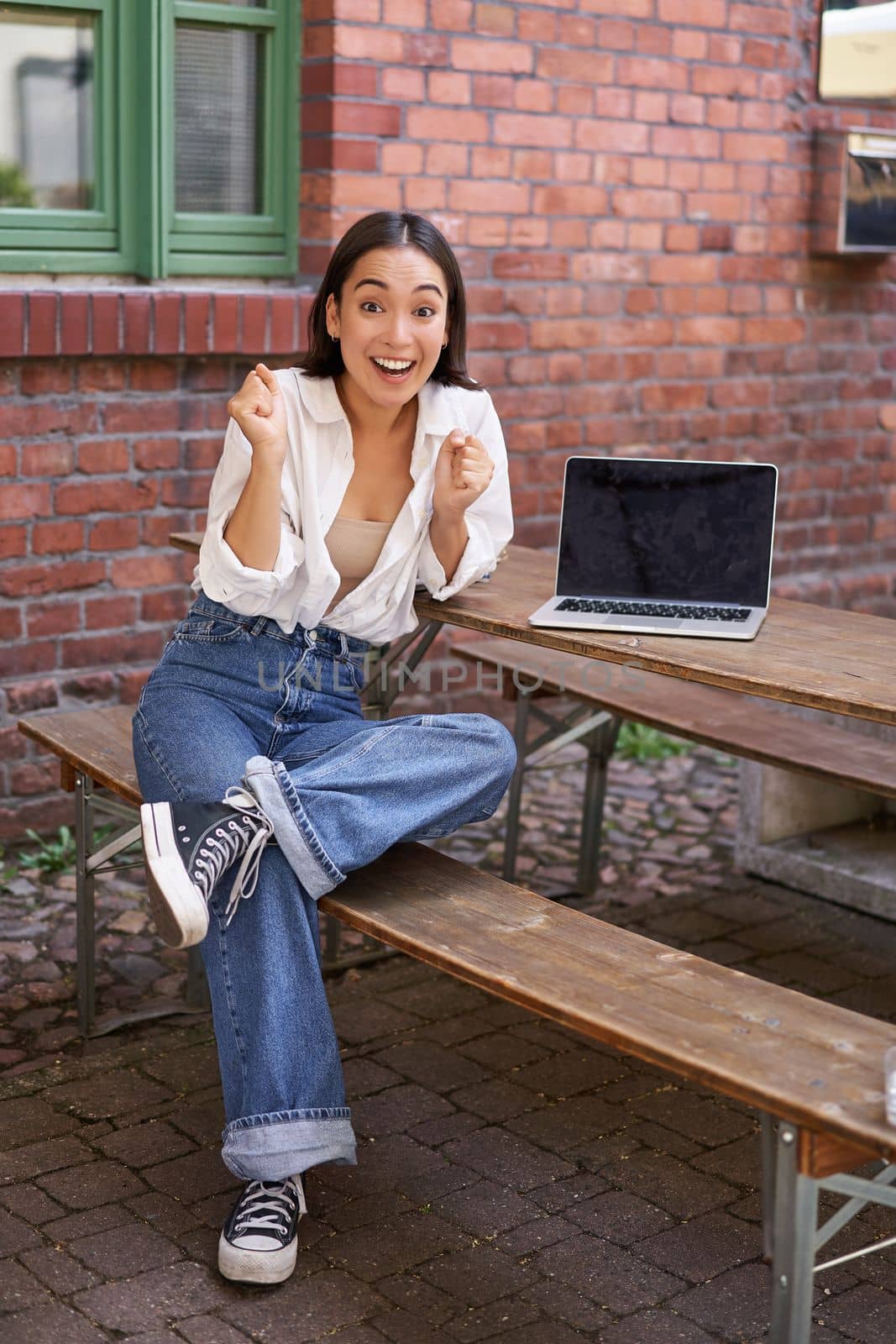 Vertical shot of enthusiastic asian girl, sits with laptop, screen is empty and blank, woman laughing and celebrating, triumphing by Benzoix