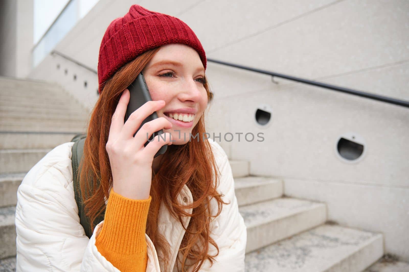 Young stylish redhead girl in red hat, sits on street and talks on mobile phone, has telephone conversation, rings her friend while relaxes outdoors by Benzoix