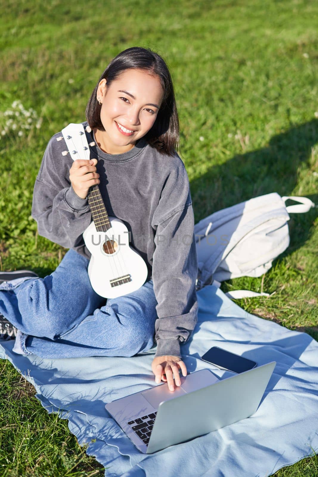 Smiling asian girl learns how to play ukulele on laptop, video chat with music teacher, sitting with instrument in park on grass by Benzoix