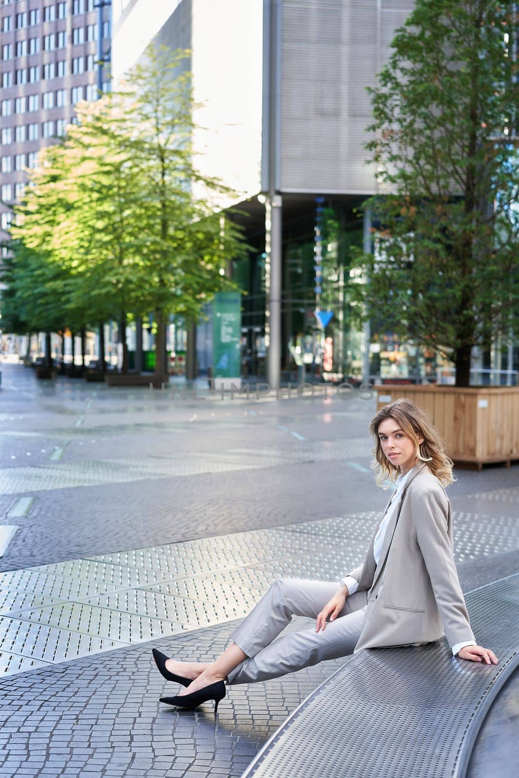 Vertical shot of beautiful businesswoman in beige suit, sitting outdoors in city centre and smiling at camera.