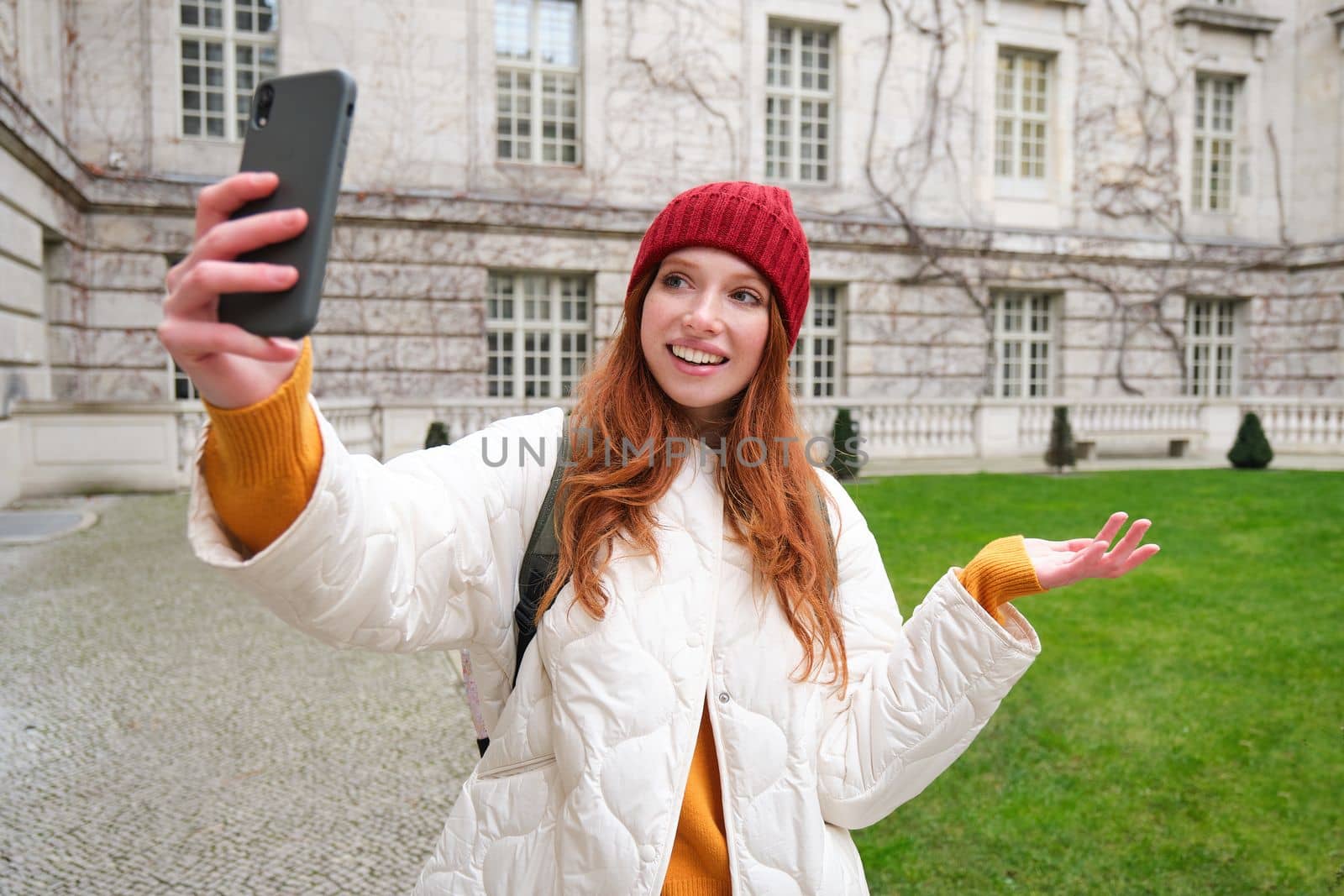 Portrait of happy girl, tourist posing near city attraction, video chats and demonstrates landmark, shows something to friend with mobile phone app.