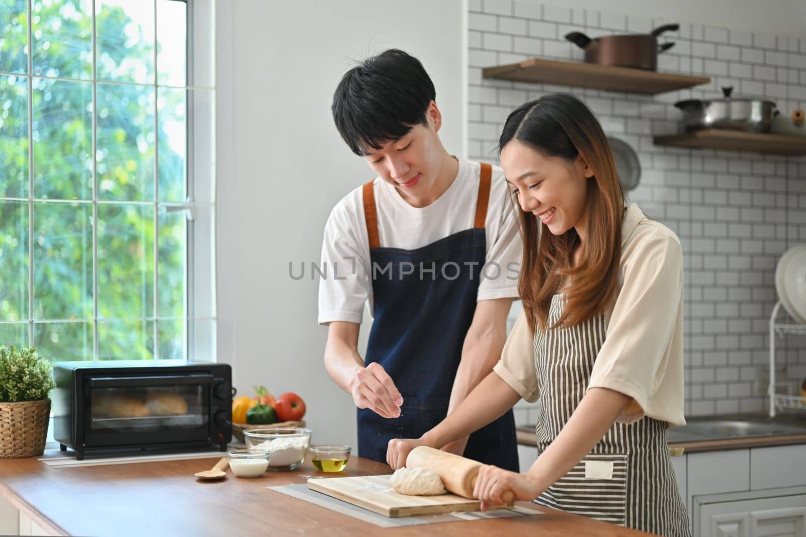 Happy young husband and wife wearing aprons preparing homemade pastry in modern kitchen interior.