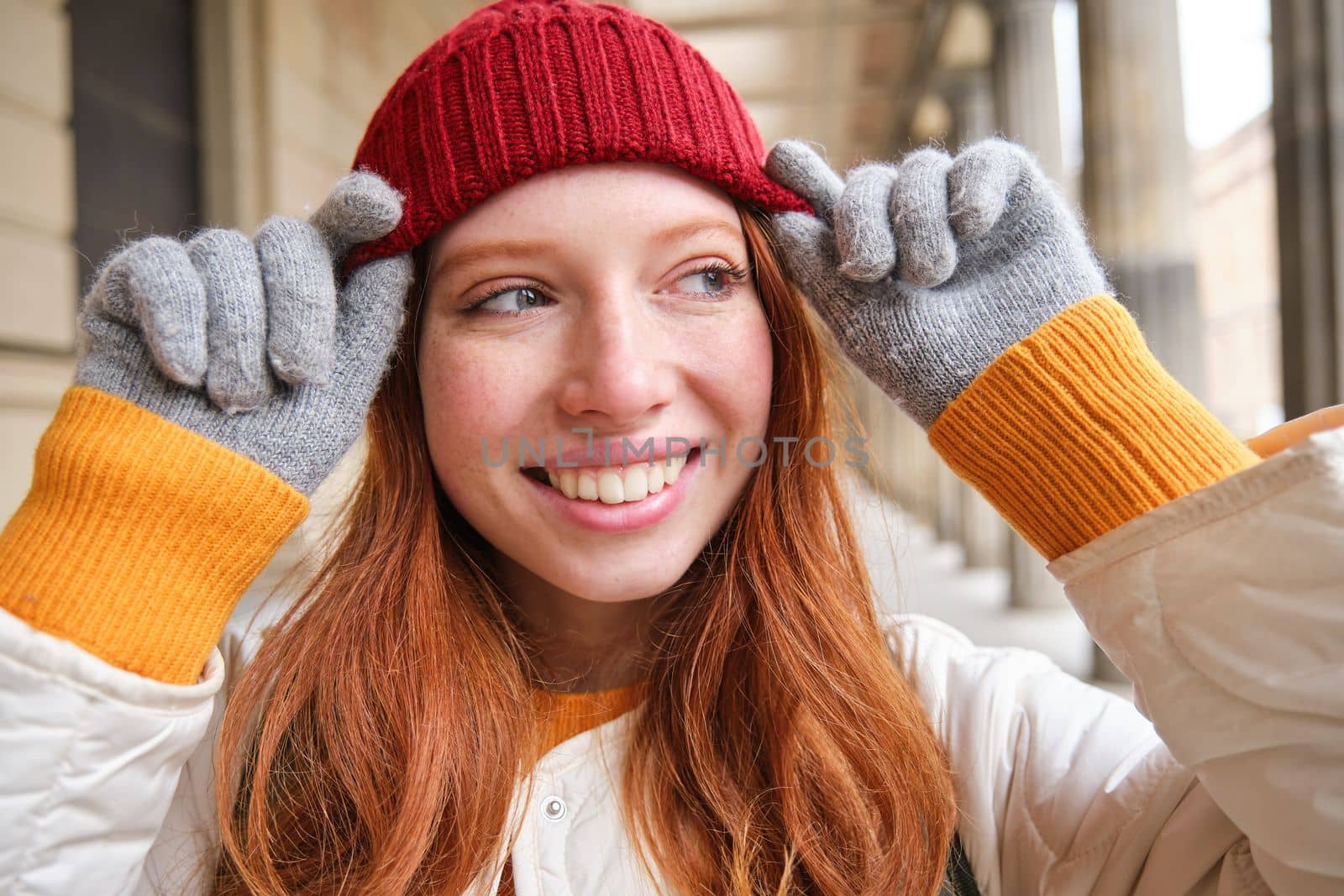 Portrait of young redhead woman in knitted hat and gloves, smiles and looks aside, walks around city in winter. Copy space