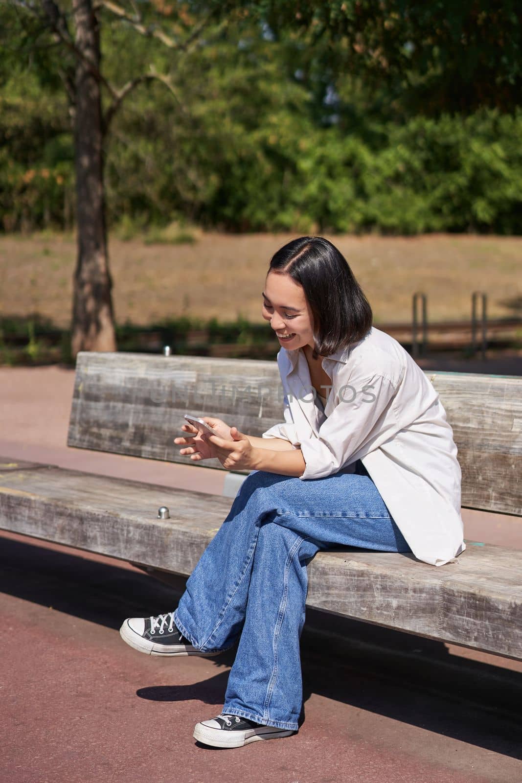 Happy smiling asian girl sitting on bench in park and browsing on social media, holding smartphone, using mobile phone app by Benzoix
